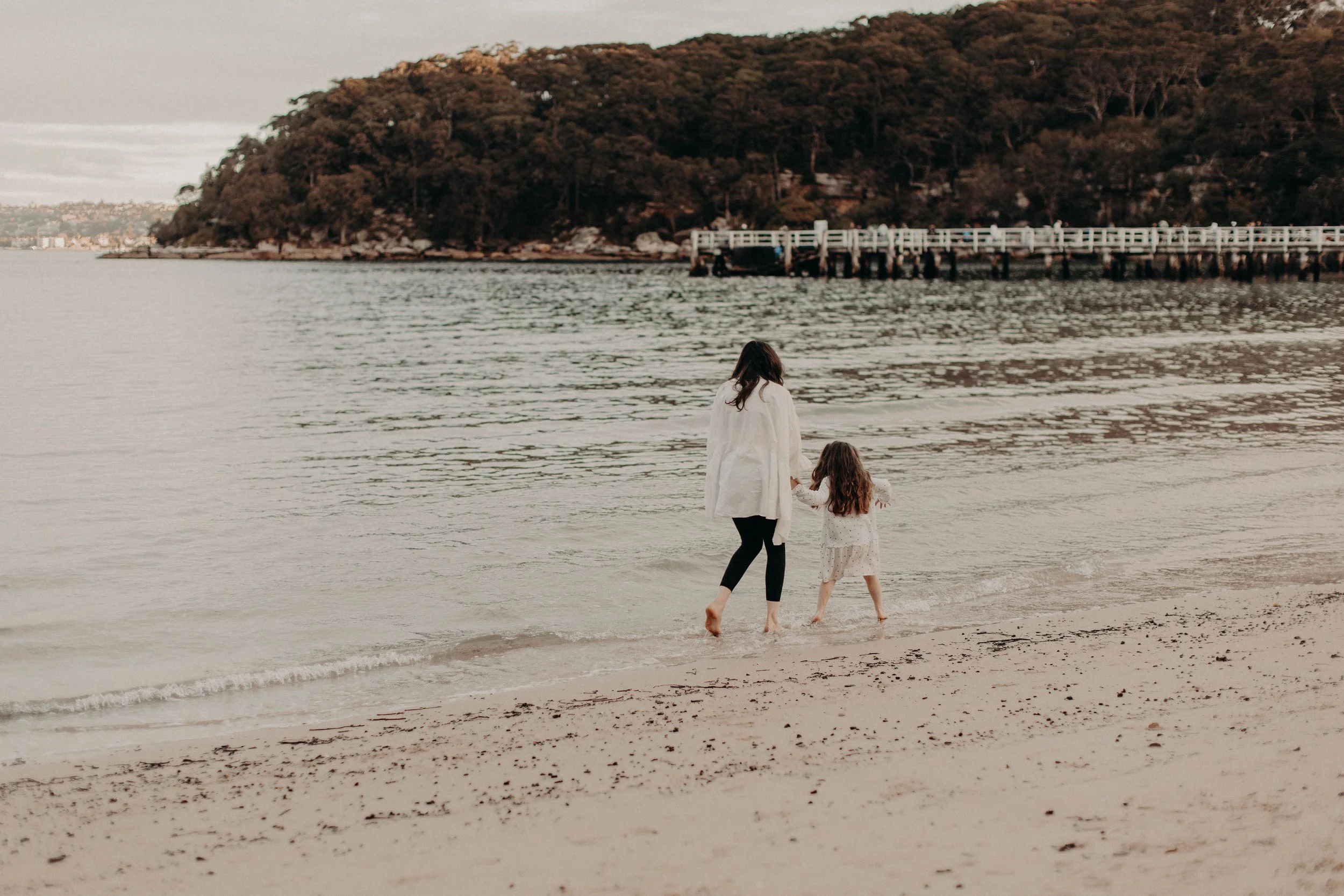 A woman and a young girl walk barefoot along the sandy shore of a body of water, with a large hill covered in trees and a pier in the background.