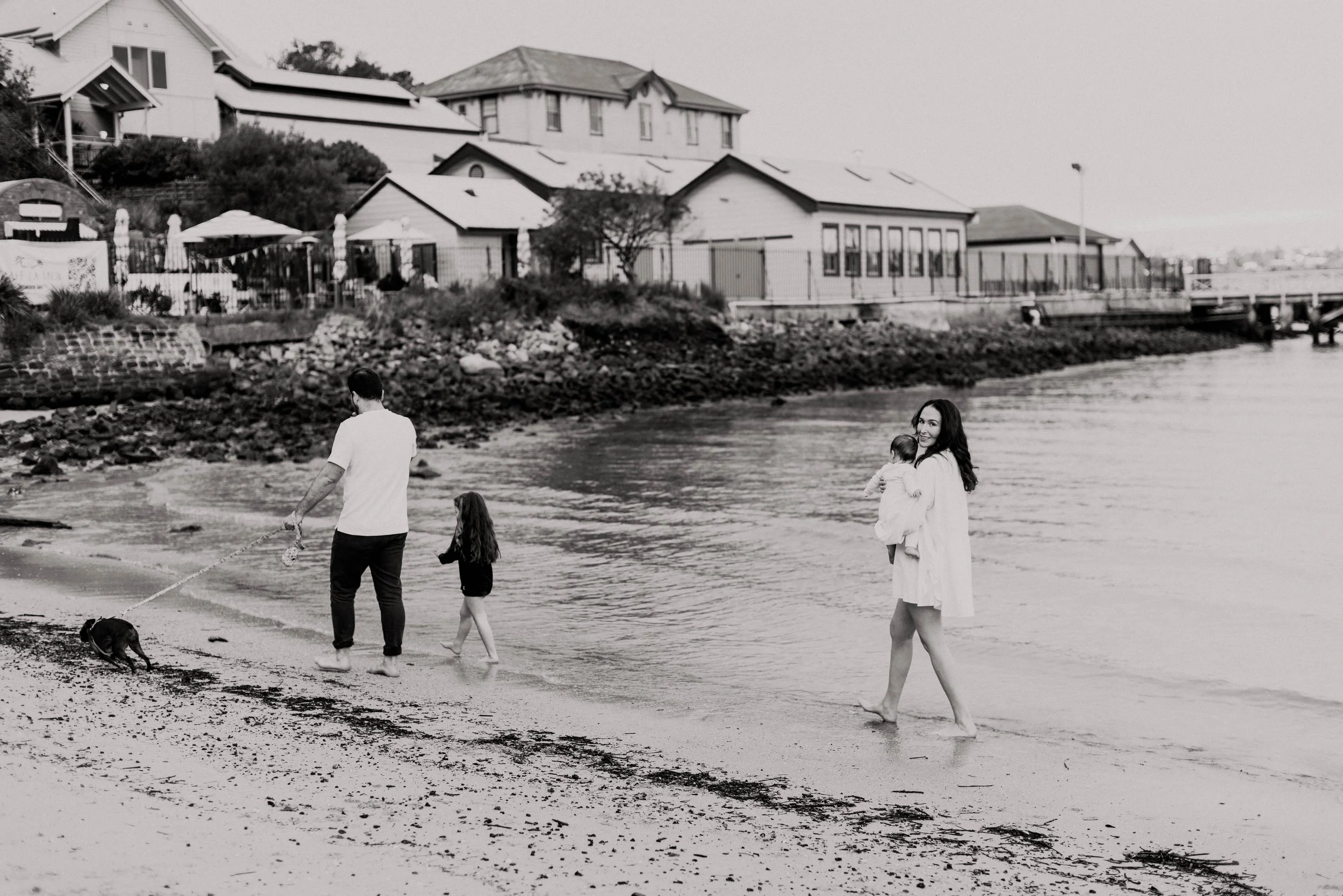 People walking along the shoreline with houses in the background, black and white photo