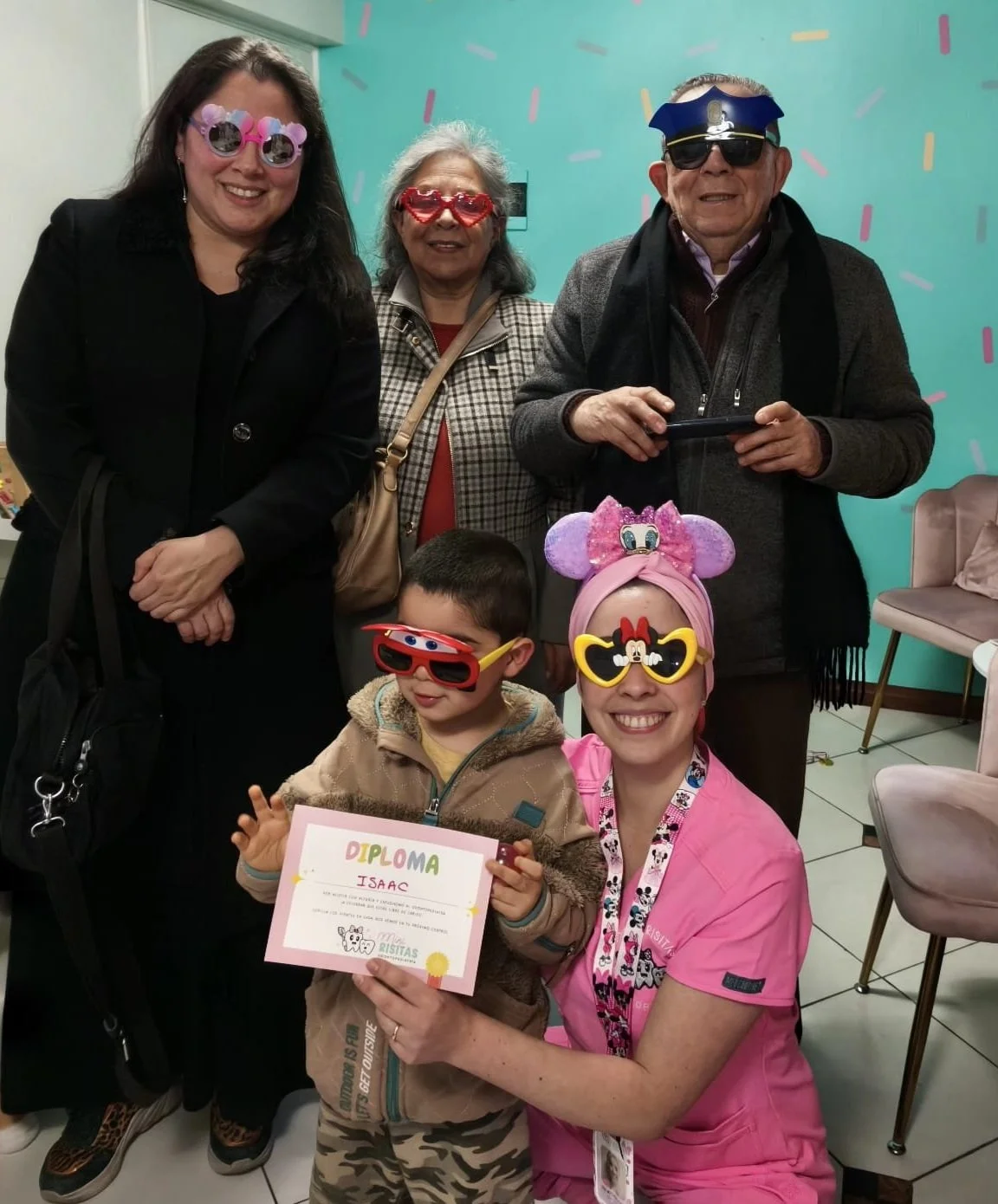 Niños y adultos en una celebración con accesorios de fiesta, algunos con gafas y disfraces. Un niño sostiene un diploma y una mujer con disfraz de Minnie Mouse sonríen juntos.