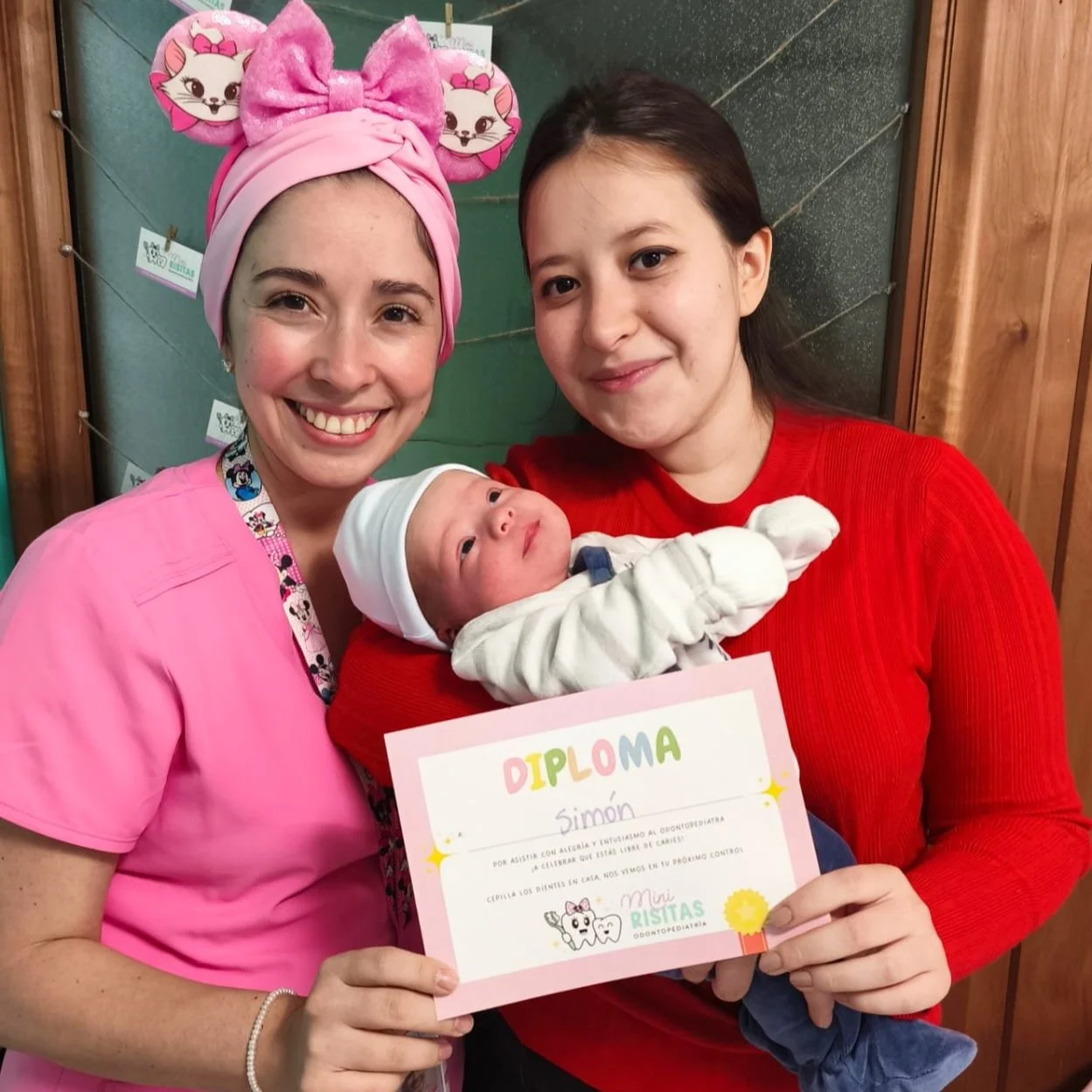 Dentista con un gorro rosa con orejas de gato, una niña con una camiseta roja y una odontopediatra con uniforme rosa, sosteniendo a un bebé con gorro blanco y diploma, en un entorno escolar.