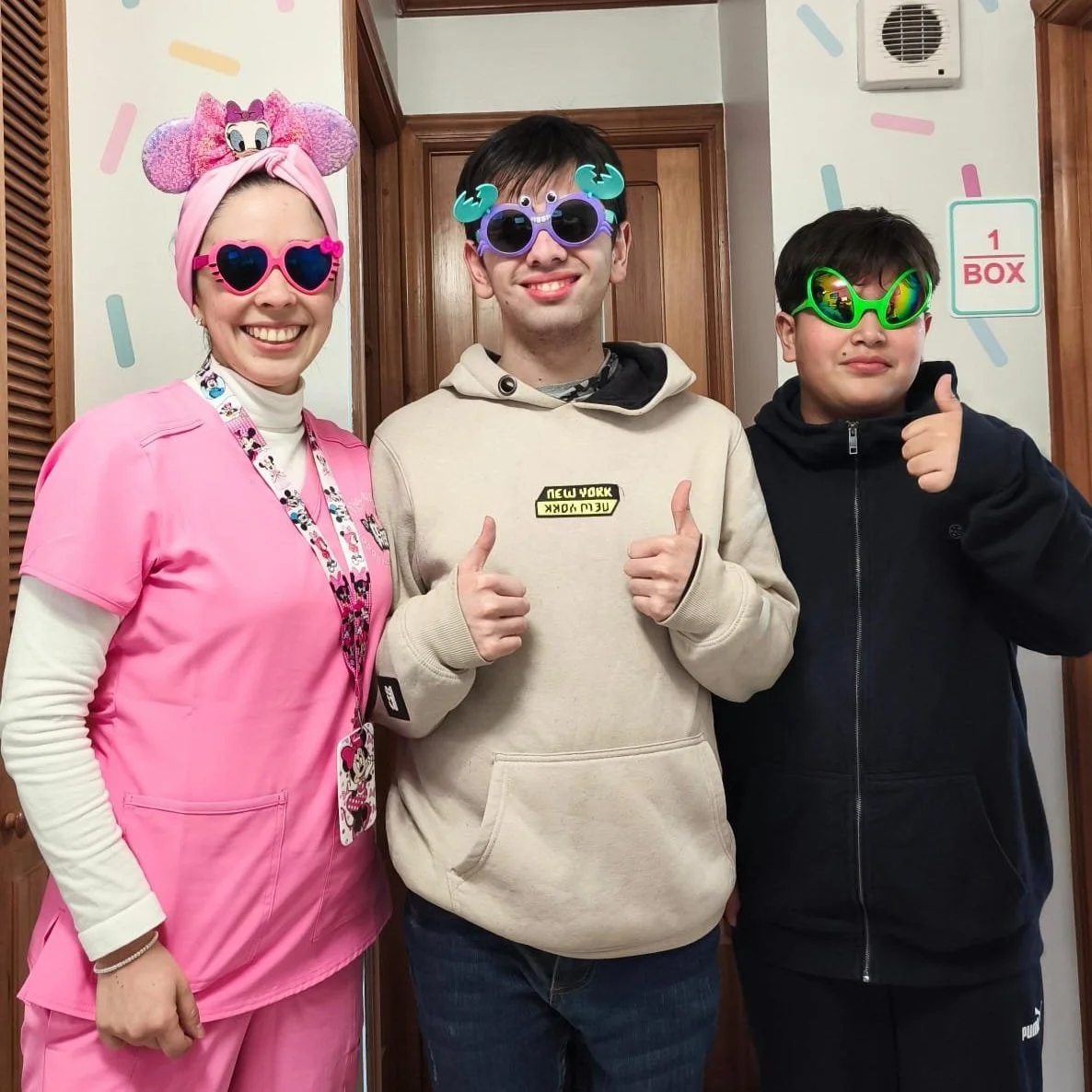 Tres personas con gafas y accesorios divertidos posando en una habitación con paredes decoradas con manchas de colores y señal de caja, sonriendo y dando mano como señal de aprobación.