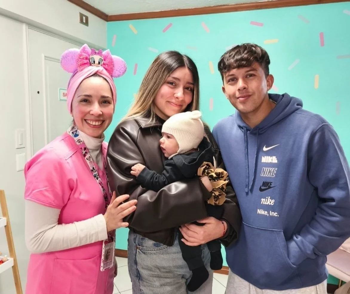 Cuatro personas posando juntas en un ambiente interior, con un fondo color turquesa con confeti. Dentista con un gorro rosa con orejas y un uniforme rosa, una joven con cabello rubio, una bebé con gorro blanco, y un joven con sudadera azul.
