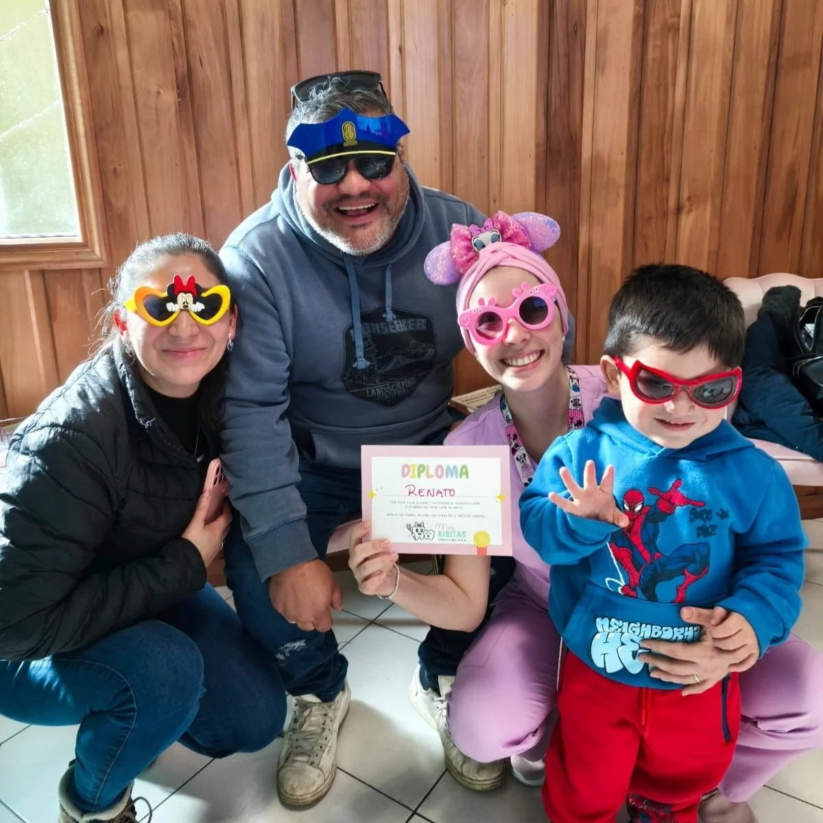 Grupo de cinco personas, incluyendo niños y adultos, con accesorios divertidos, celebrando y sosteniendo un diploma con el nombre Renato, en un ambiente de madera, todos sonriendo.