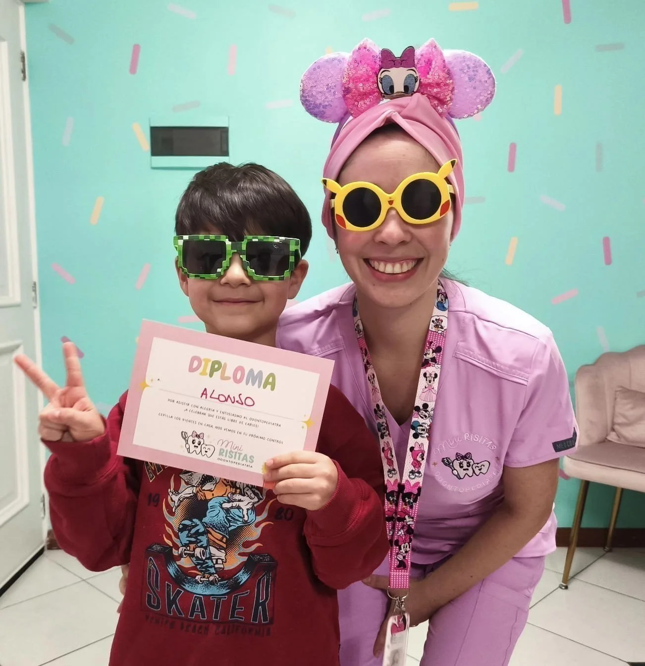 Niño con gafas de sol verdes y camiseta roja, sosteniendo un diploma de reconocimiento, junto a una mujer con gafas de sol amarillas, bigote de Minnie Mouse y gorro de Minnie Mouse rosa, en un fondo color aqua con confeti de colores.