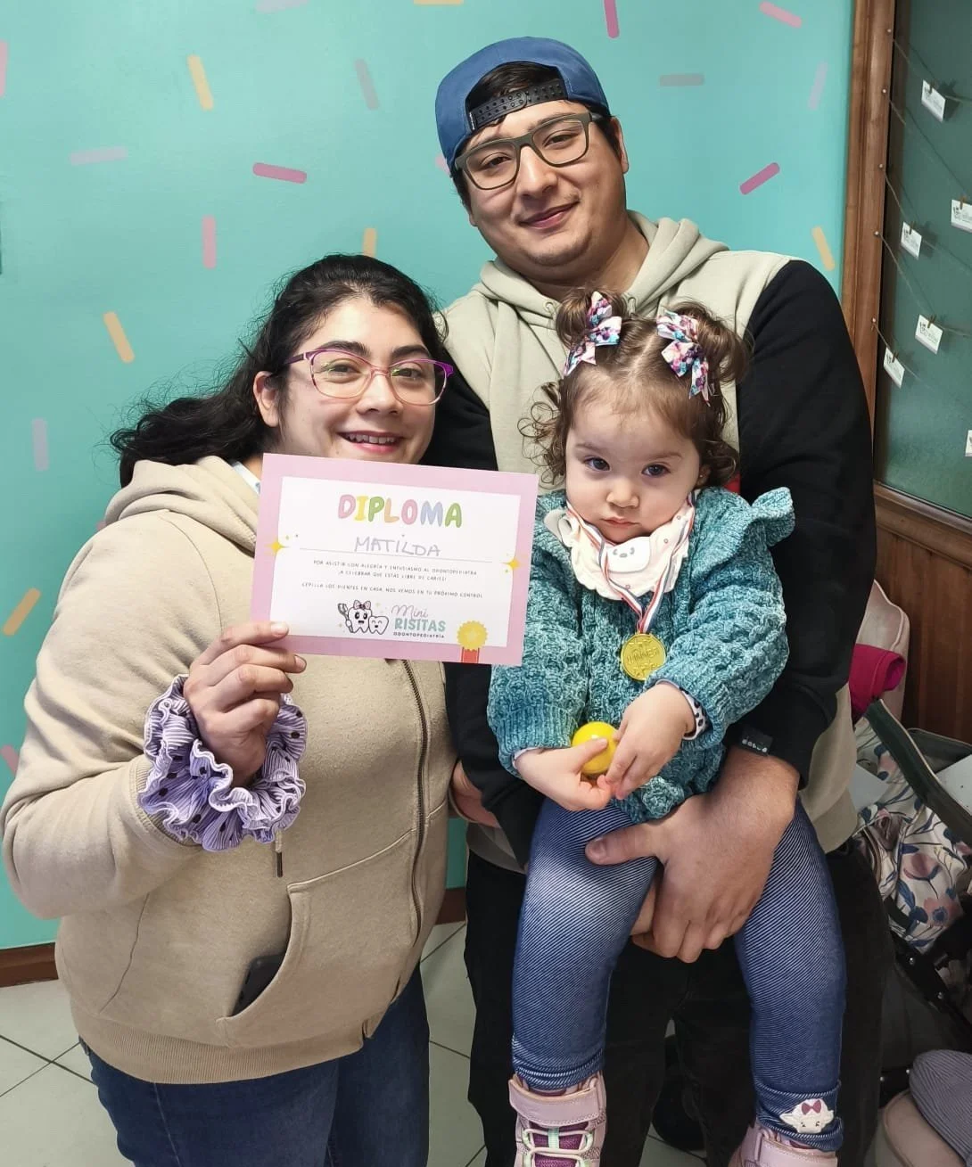 Una mujer, un hombre y una niña con una medalla, celebrando una graduación, sosteniendo un diploma, en un fondo color aqua con confeti decorativo.
