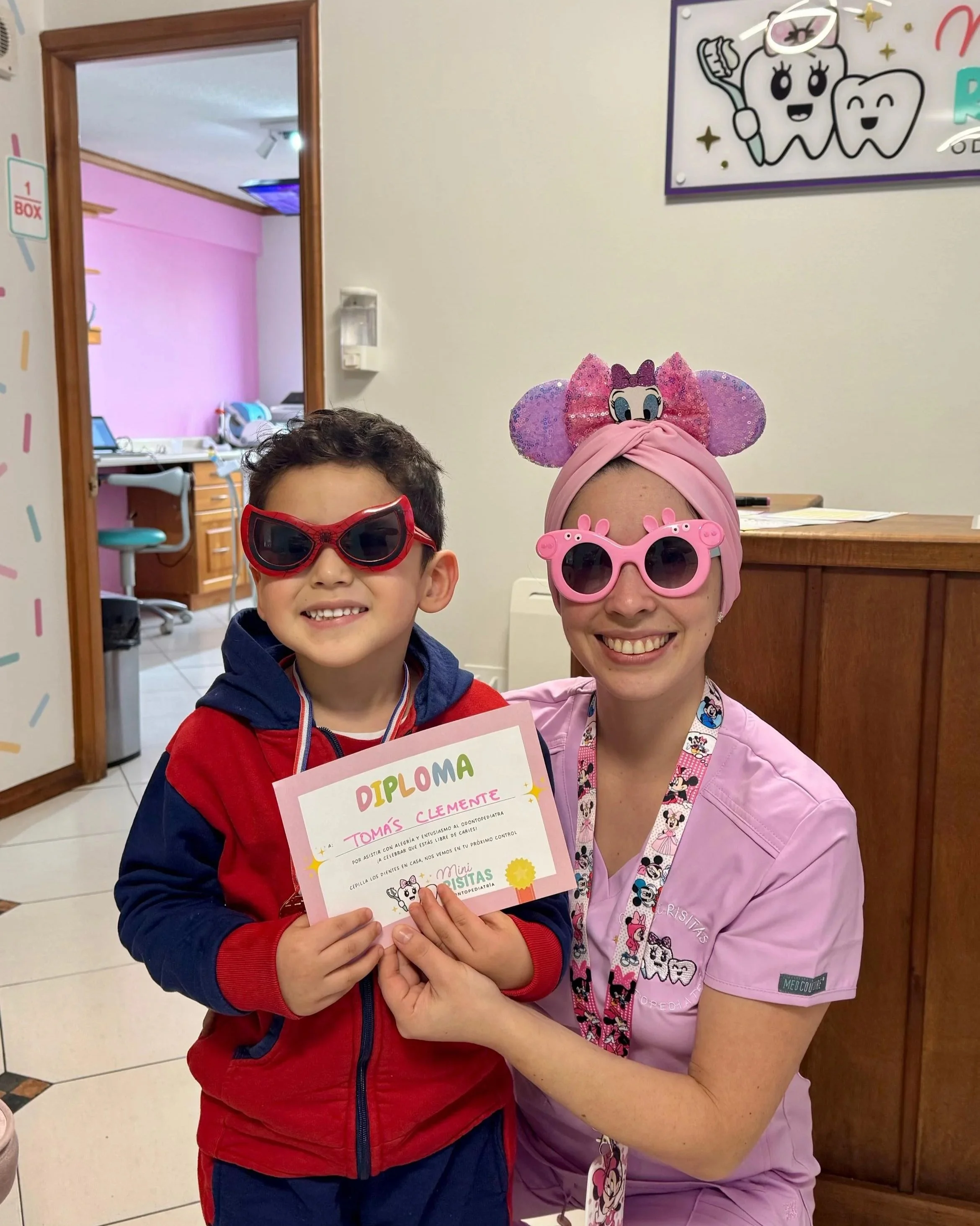 Niño y mujer con gafas de sol y sombreros divertidos en una clínica dental, sosteniendo un diploma de reconocimiento, sonriendo. La clínica tiene decoraciones infantiles y un ambiente alegre.
