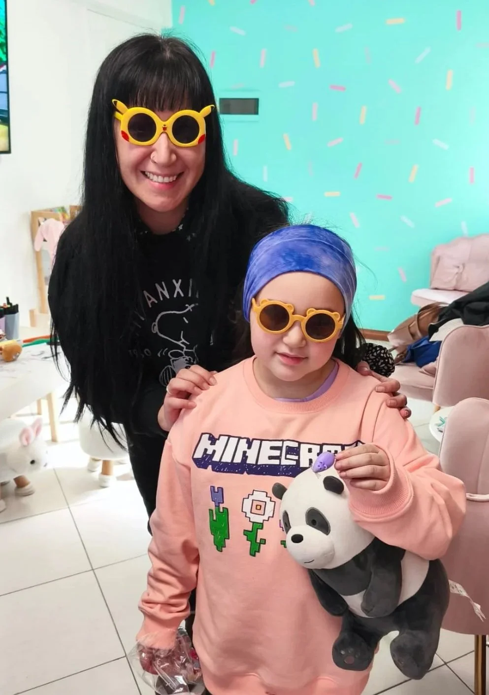 Una mujer y una niña con gafas de sol amarillas y sonrientes, la mujer lleva gafas con forma de Pikachu y la niña con una bufanda azul. La niña sostiene un peluche de panda. Están en un ambiente decorado con colores brillantes y decoraciones festivas