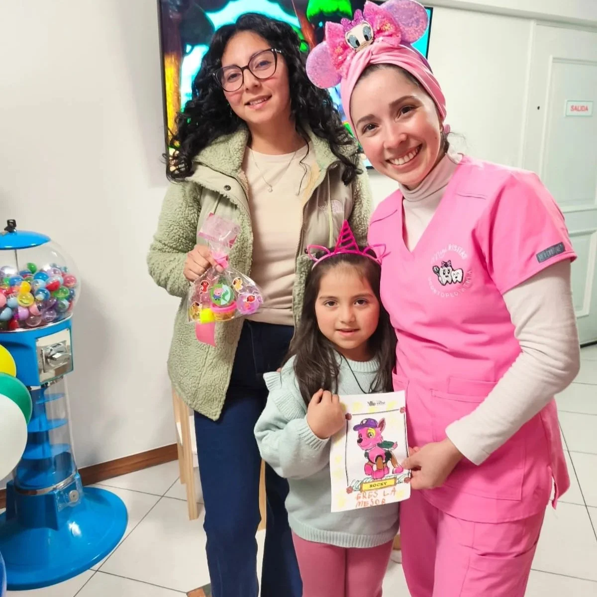 Niña con orejas de unicornio rosa recibe premio y feliz junto a dos mujeres, una con bata rosa y otra con chaqueta verde, en una celebración infantil.