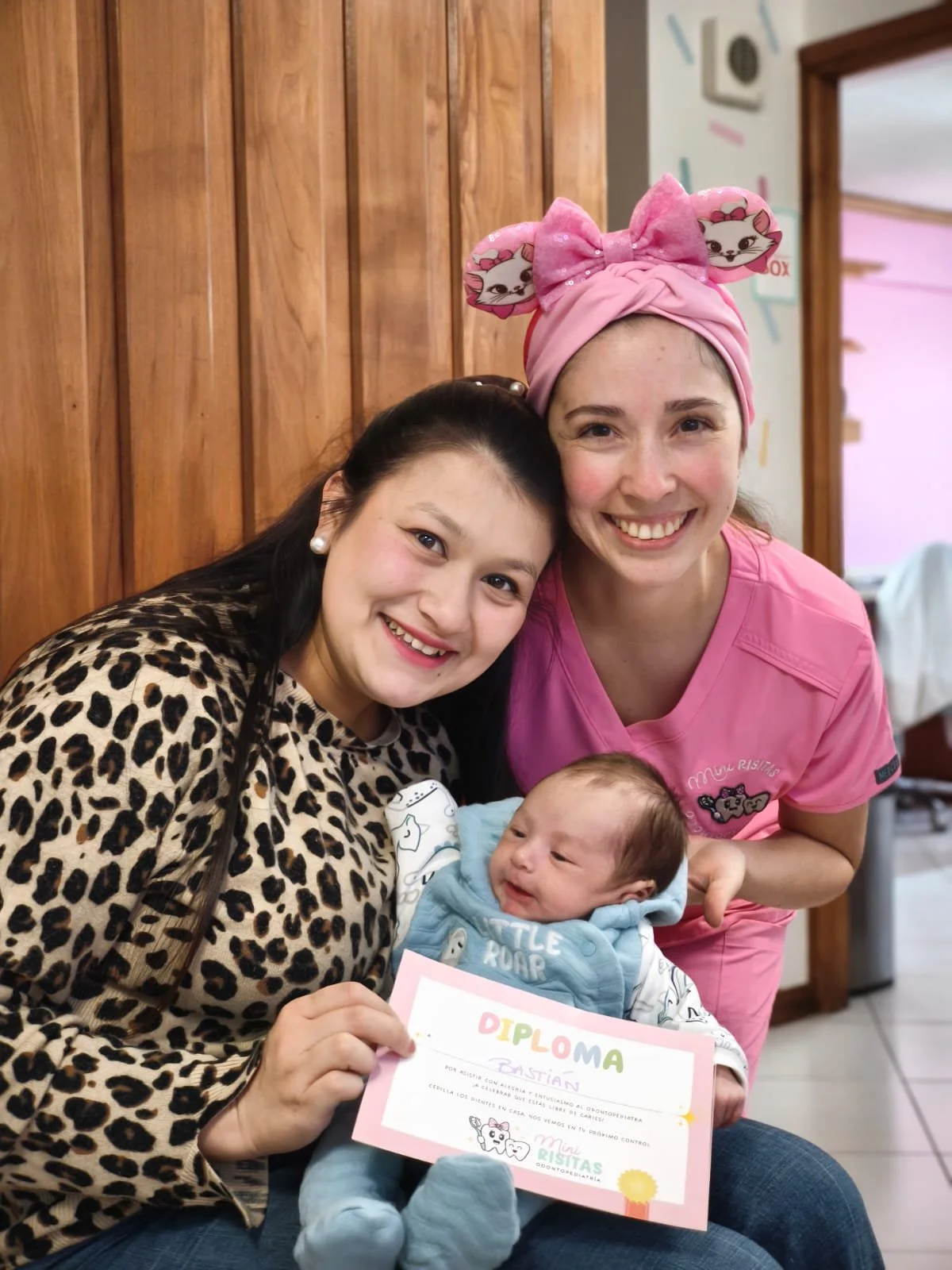 Mujer con gorro rosa y mujer con blusa de animal print sosteniendo a un bebé que tiene un diploma, en un ambiente de clínica u hospital, celebrando un logro o graduación.
