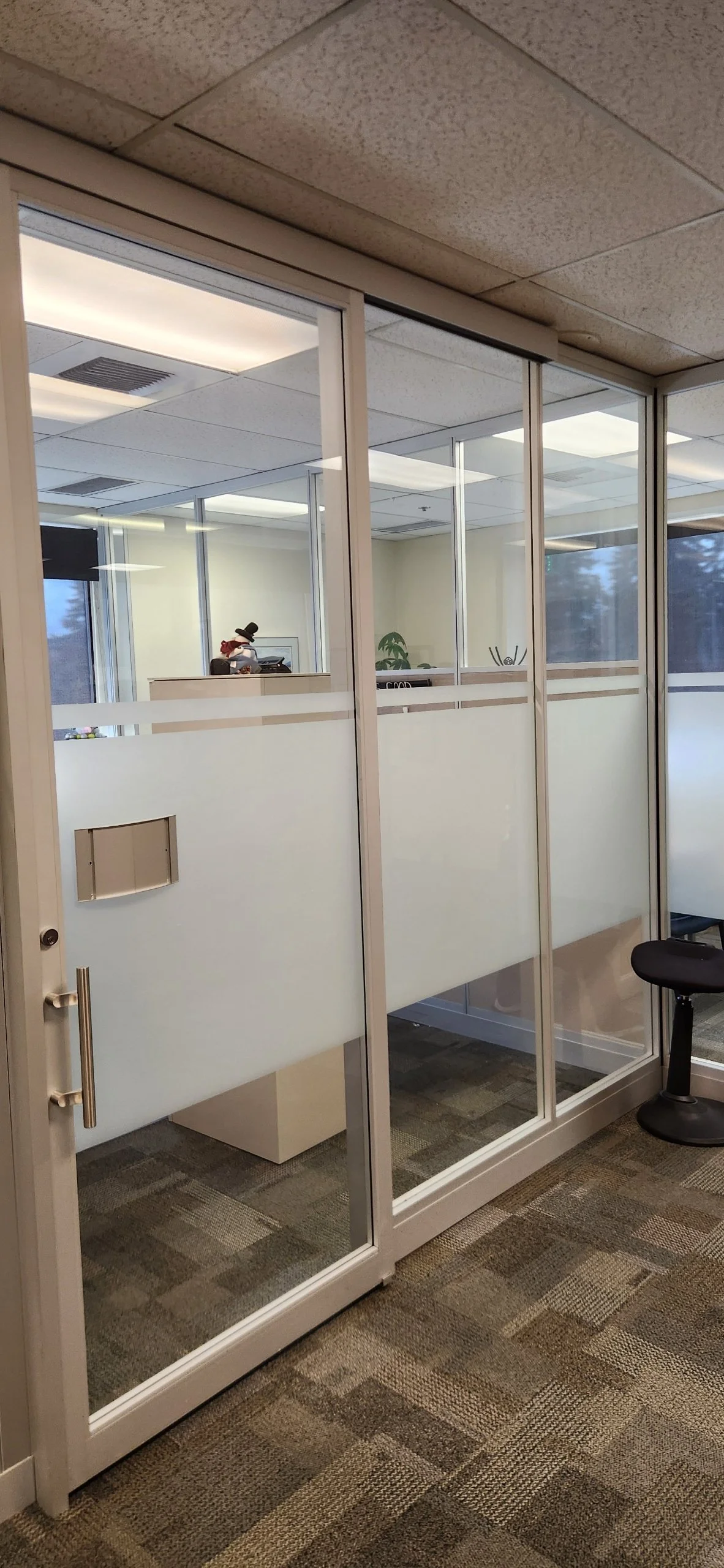 View through glass-walled office door showing an office interior with a figure in a snowman costume at a desk, office plants, and ceiling lights.