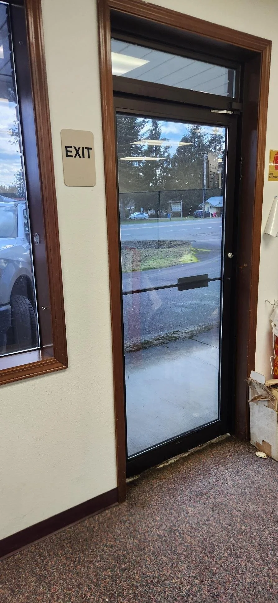 The image shows a glass exit door with a view of a parking lot and trees outside. There is an exit sign on the wall next to the door and some debris or packaging on the bottom right corner. The floor is carpeted.