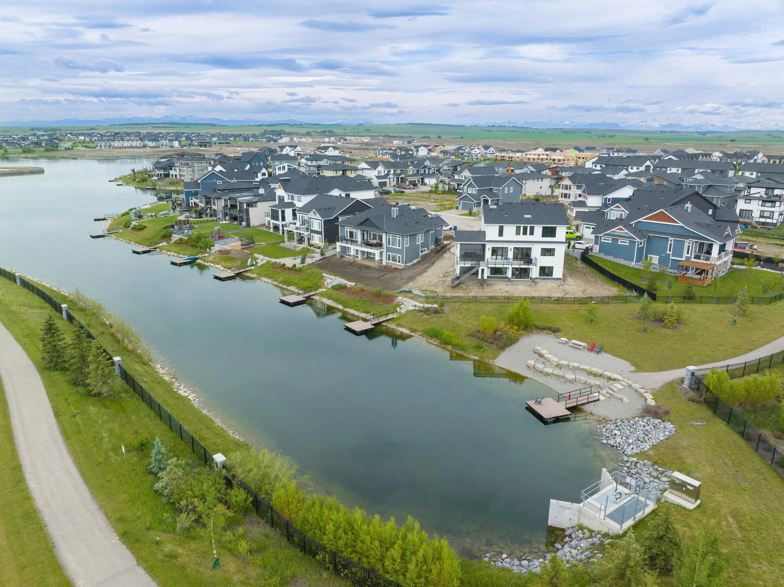 Aerial view of a residential neighborhood with modern houses by a river, surrounded by green grass and trees, under a partly cloudy sky.