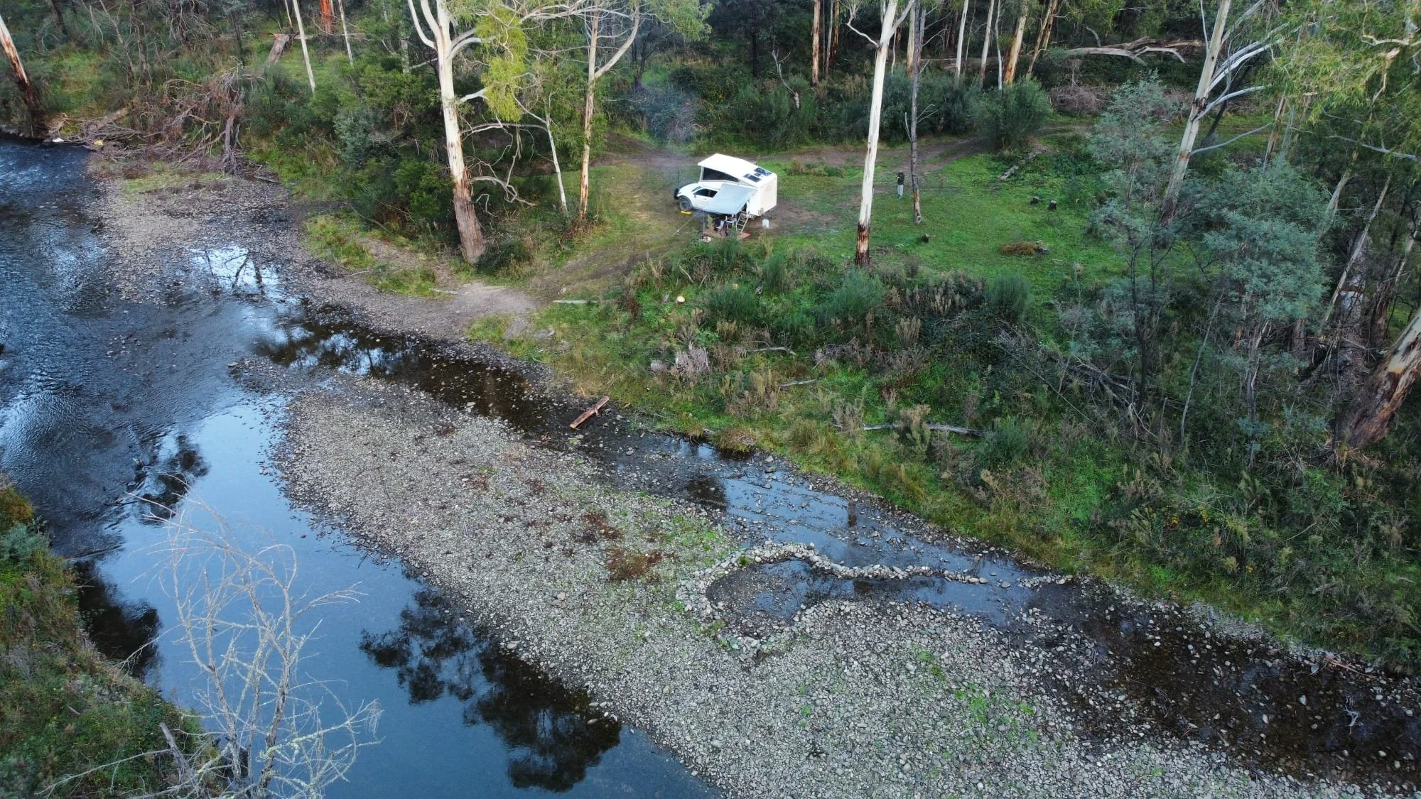 Aerial view of a riverside campsite with a white camper trailer and vehicle parked on grass near trees, with calm river water reflecting the sky and surrounding trees.