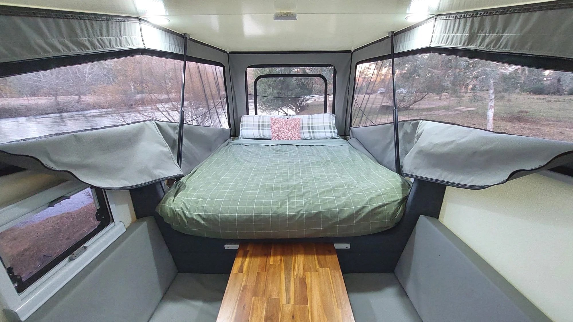 Interior view of a camper trailer with a bed, pillows, and windows showing outdoor trees and a river.