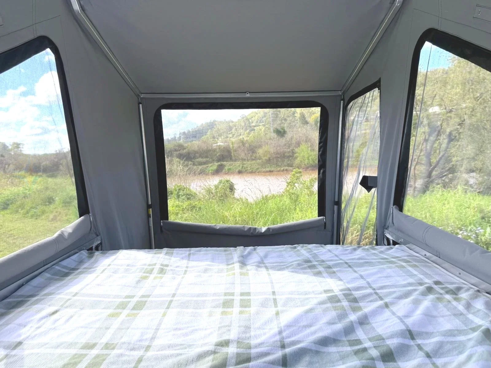 Interior of a camping trailer with a bed covered in a plaid sheet and windows showing greenery and a river outside.