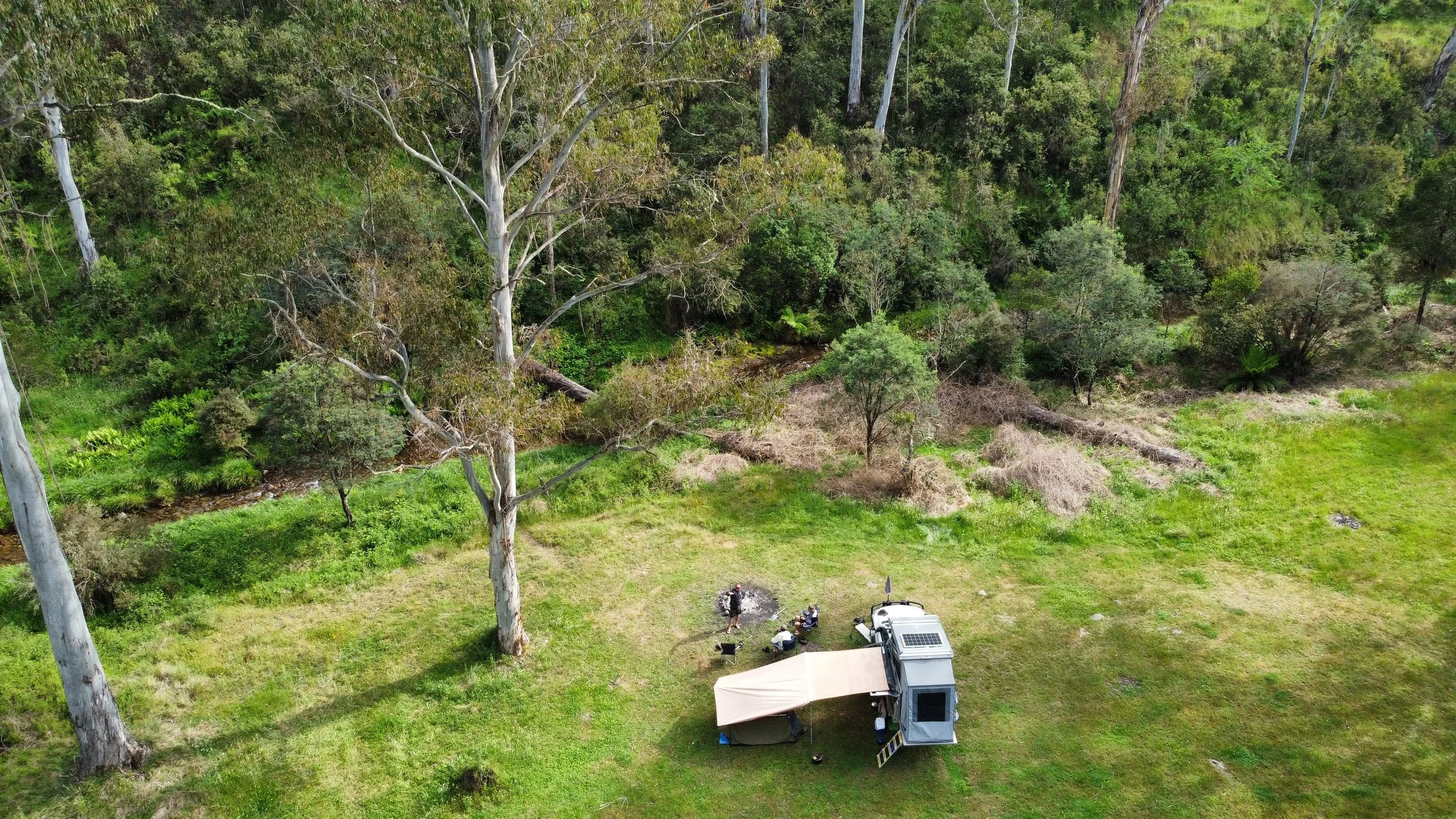 Aerial view of a campsite with a trailer, two canopies, and multiple people near a small fire surrounded by tall trees in a green forest.