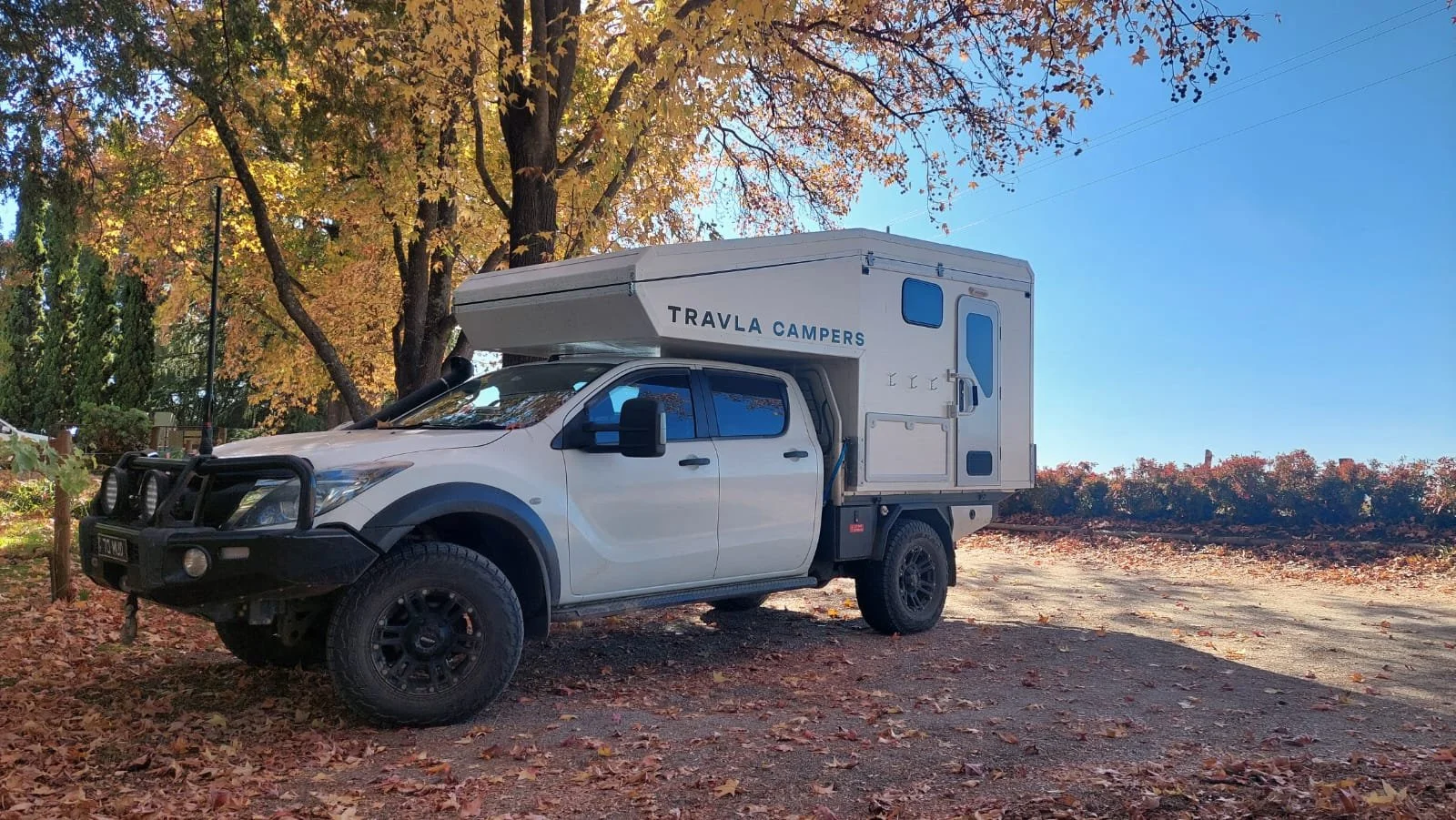 A white off-road vehicle with a camper attachment labeled "TRAVLA CAMPERS" parked on a leaf-covered trail under trees with autumn foliage.