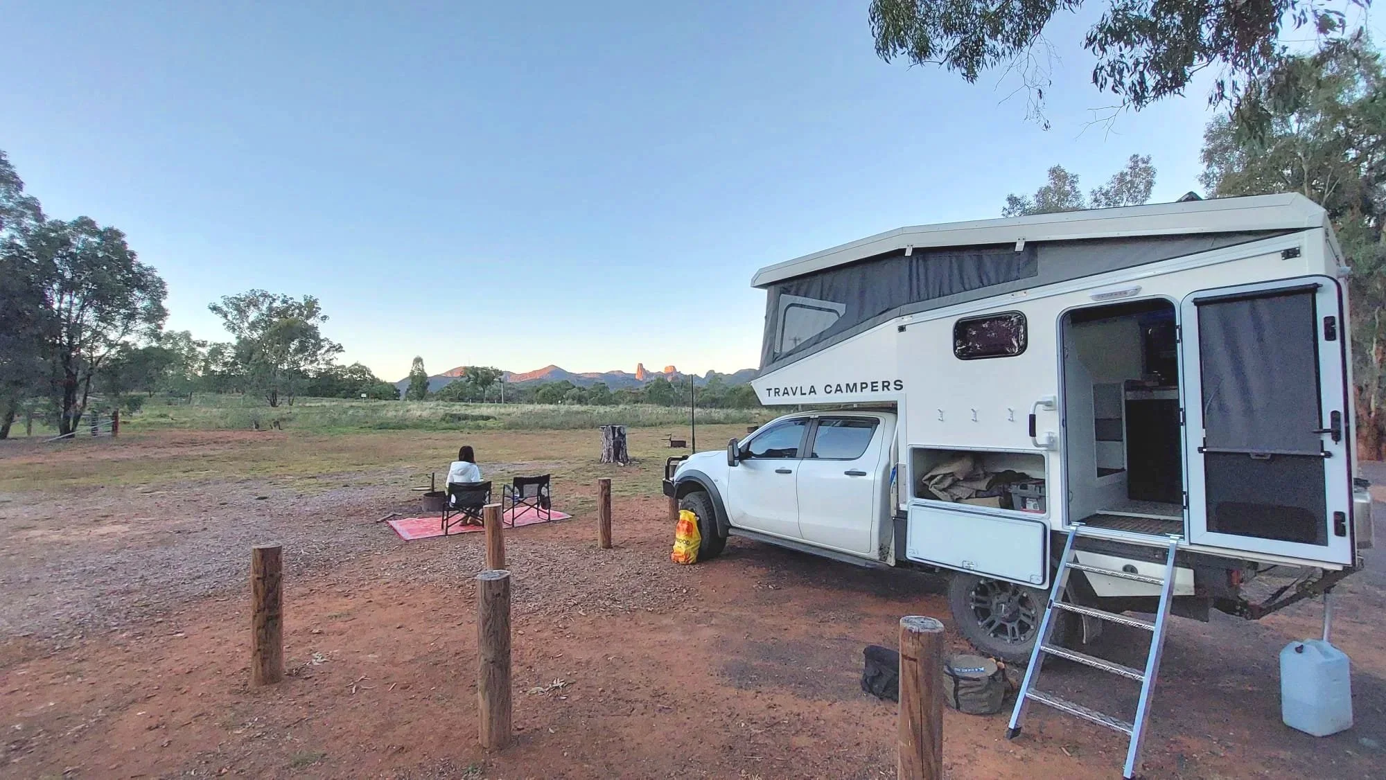 A white camper truck with a pop-up roof, parked on a dirt clearing near grass and trees, with two people sitting on chairs next to it, mountains in the background at sunset.