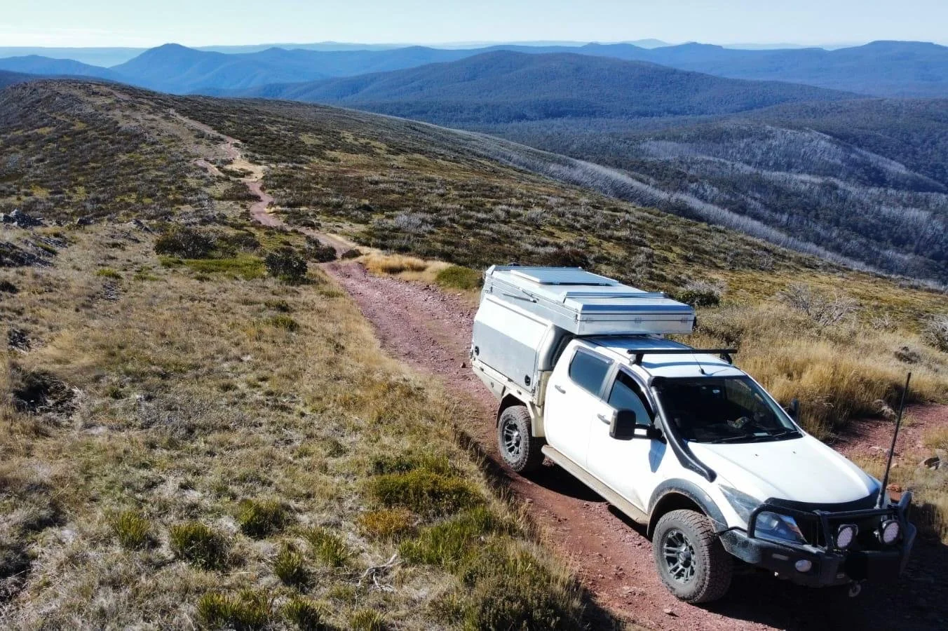 A white off-road camper vehicle on a dirt trail in a mountainous landscape with rolling hills and distant mountains.