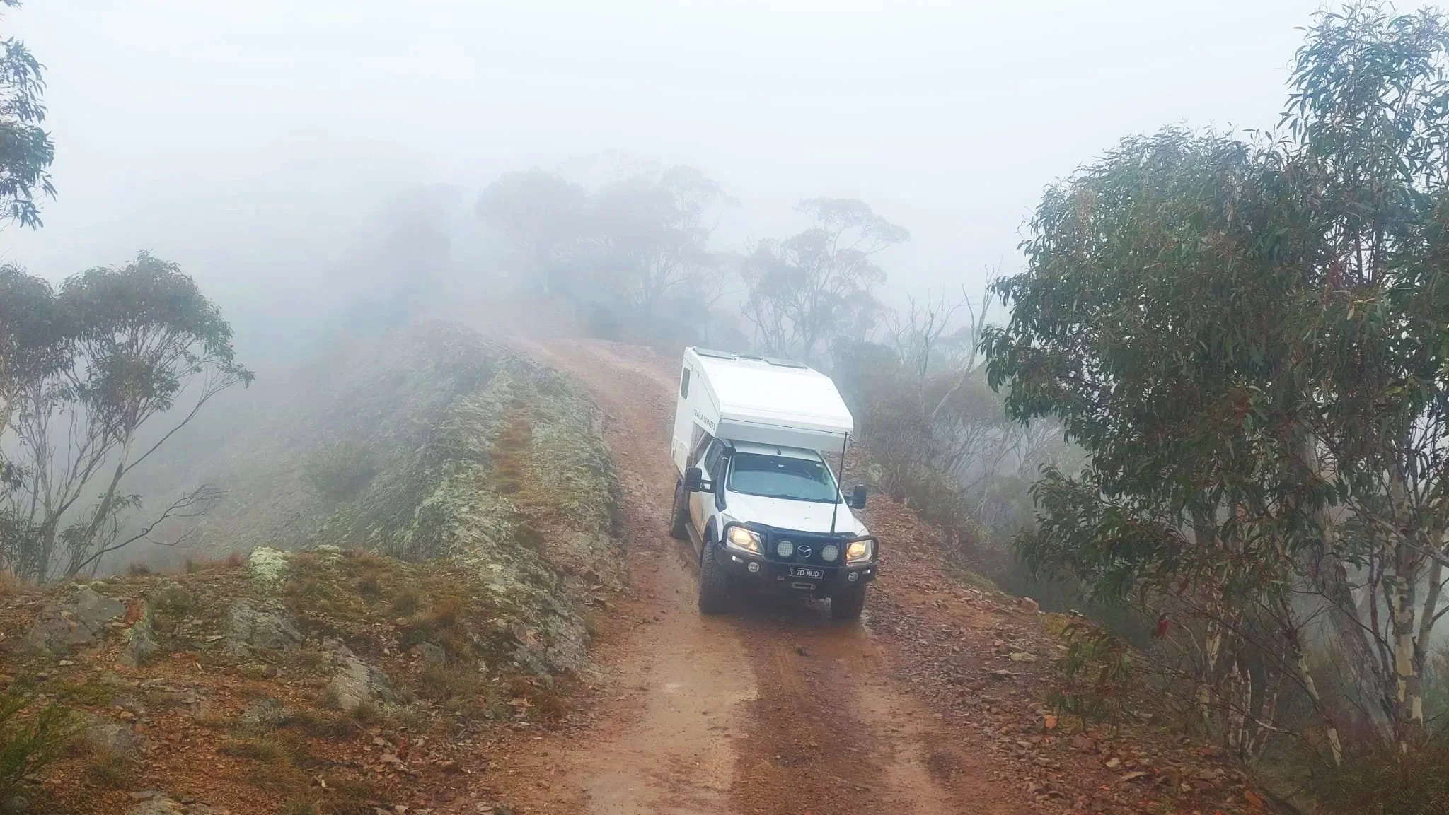A white campervan driving on a narrow, foggy, dirt mountain trail surrounded by trees.