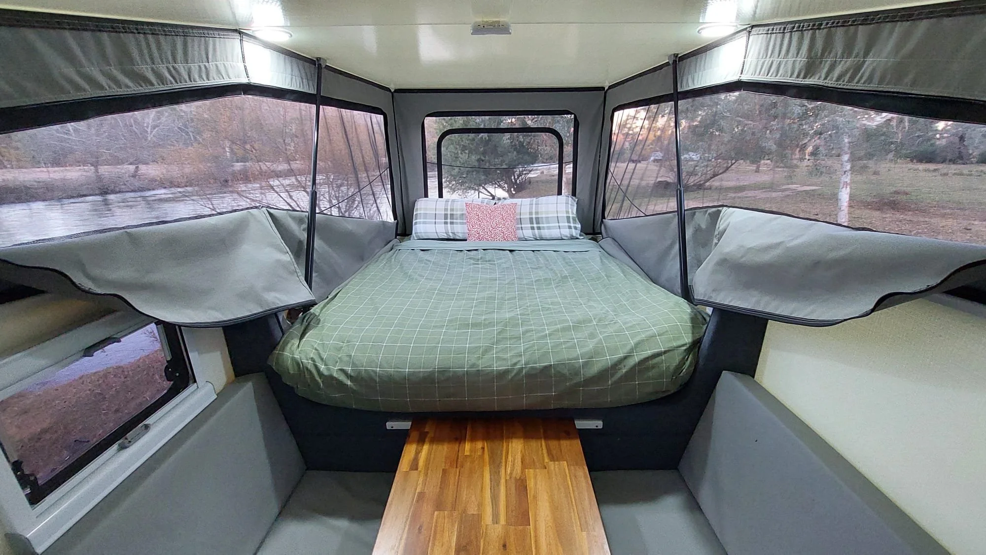 Interior of a camper with a bed, side windows, and a small wooden step leading to the bed, overlooking trees and a river outside.