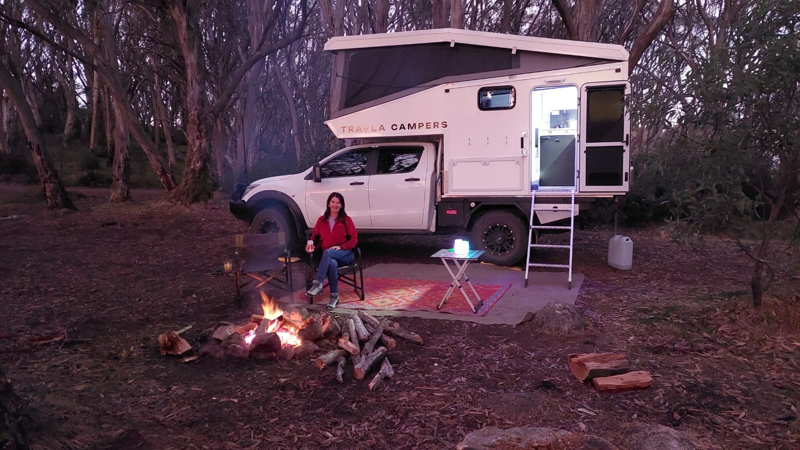 A woman sitting in a camping chair next to a campfire in front of a truck that has a travel camper attached, in a wooded area during dusk or early evening.