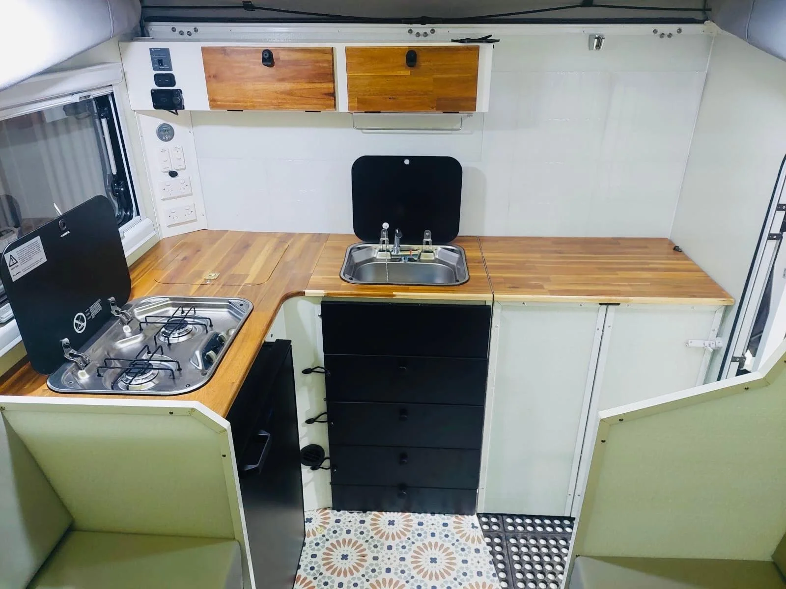 Compact kitchen inside a camper or RV with a two-burner stove, a small sink, wooden countertops, white and wood cabinets, and a patterned floor mat.