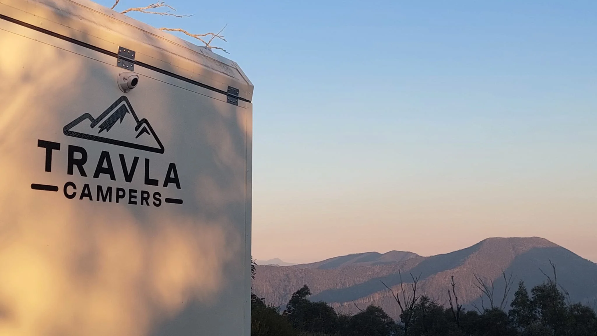 A camper trailer with the logo and name 'Travla Campers' on the side, outdoors with mountains and a clear sky in the background.