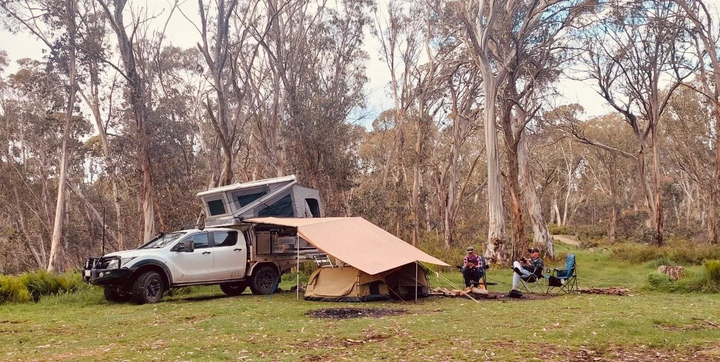 A camping scene in a forest featuring a silver pickup truck with a rooftop tent, a small tent with a canopy, portable chairs, and two people sitting and standing, surrounded by tall trees.