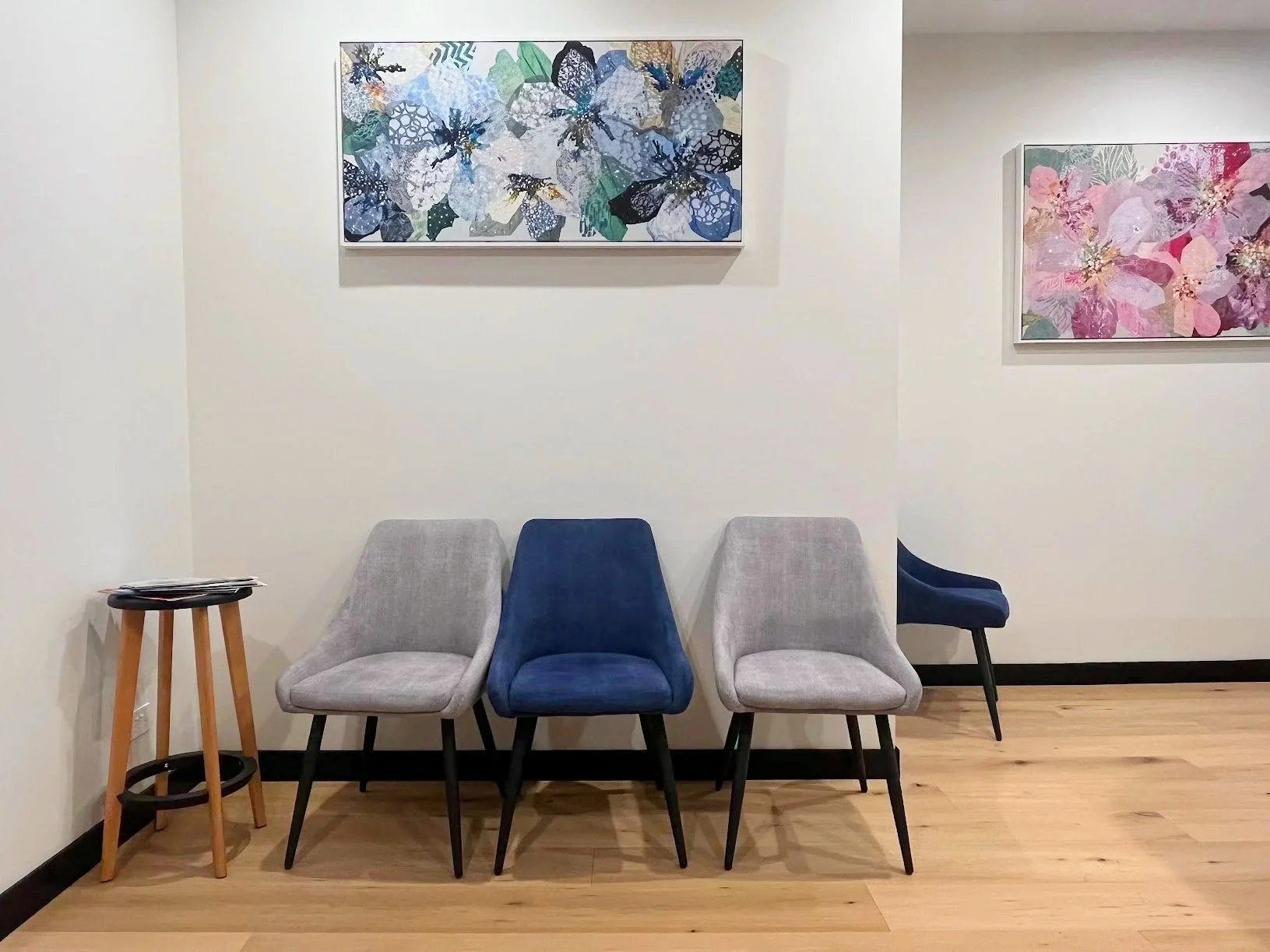 A row of four chairs in a waiting area with colorful floral paintings hanging on a white wall behind them. There is a small wooden table with some papers on it to the left.