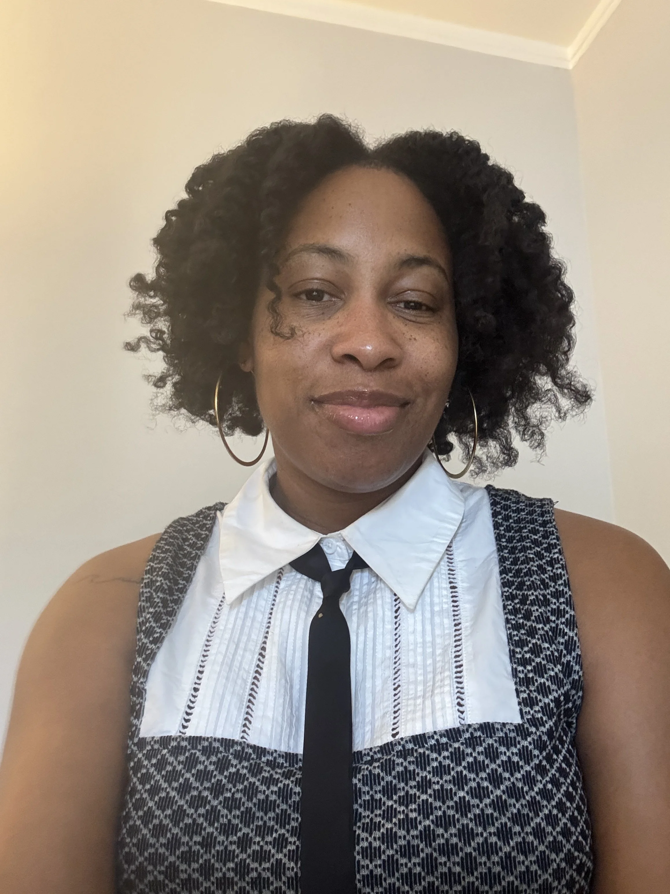 Woman with dark black curly hair and dark brown skin, wearing gold hoop earrings, a white collard top with a black and white pattern and black tie.