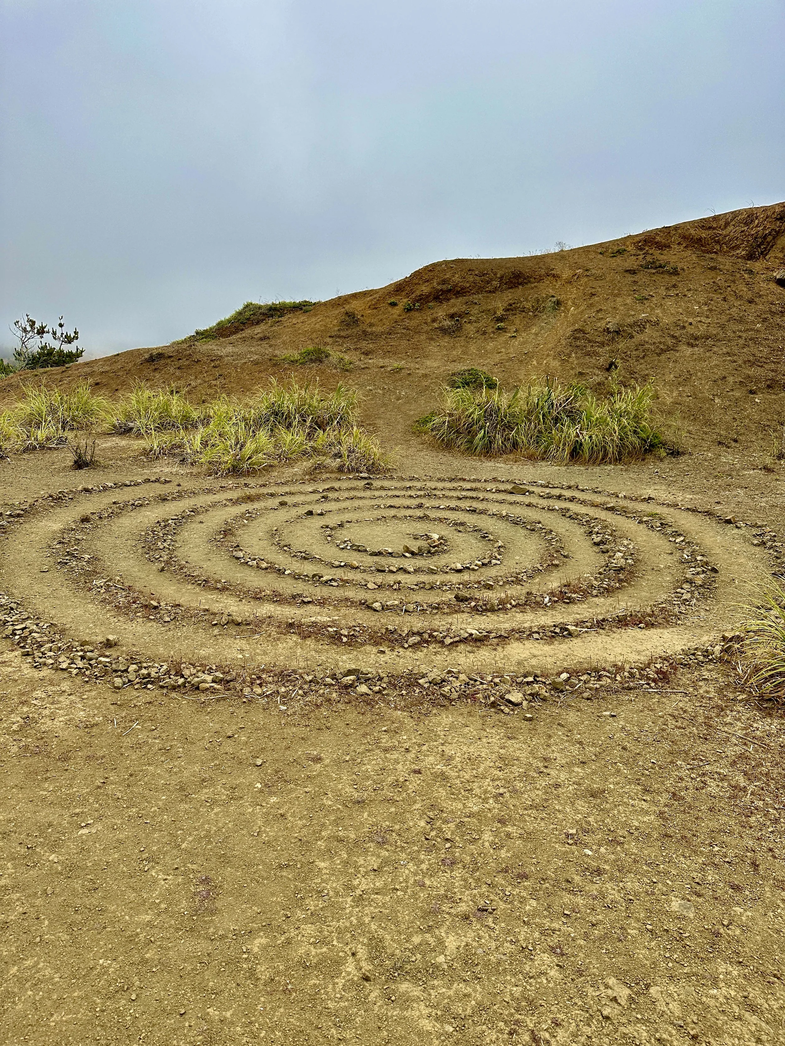 A labyrinth created in a dirt path in a hilly area with patches of grass and small plants, under a gray sky.