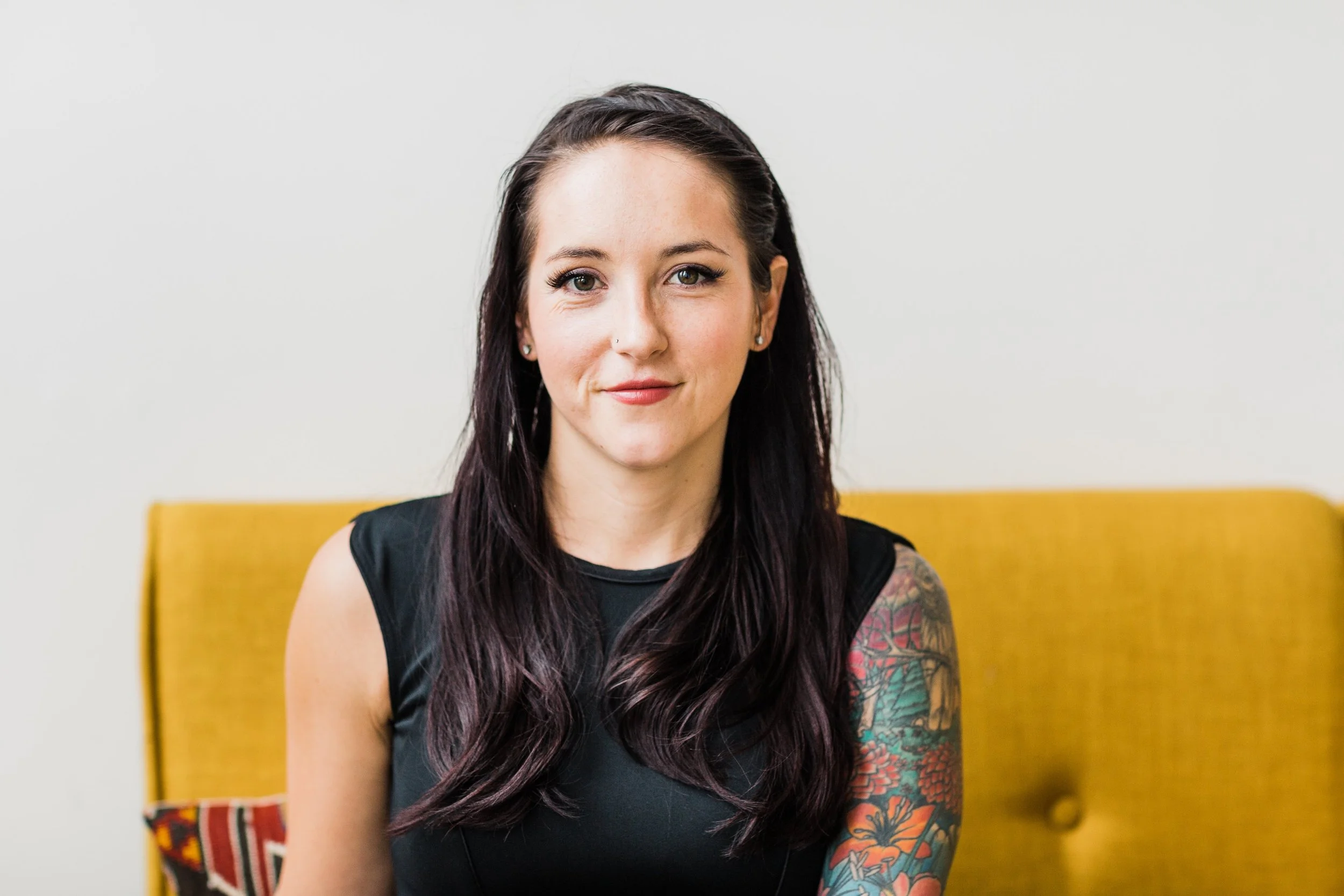 A woman with long dark hair, visible earrings, and a tattoo on her arm, sitting on a yellow chair against a plain white background.