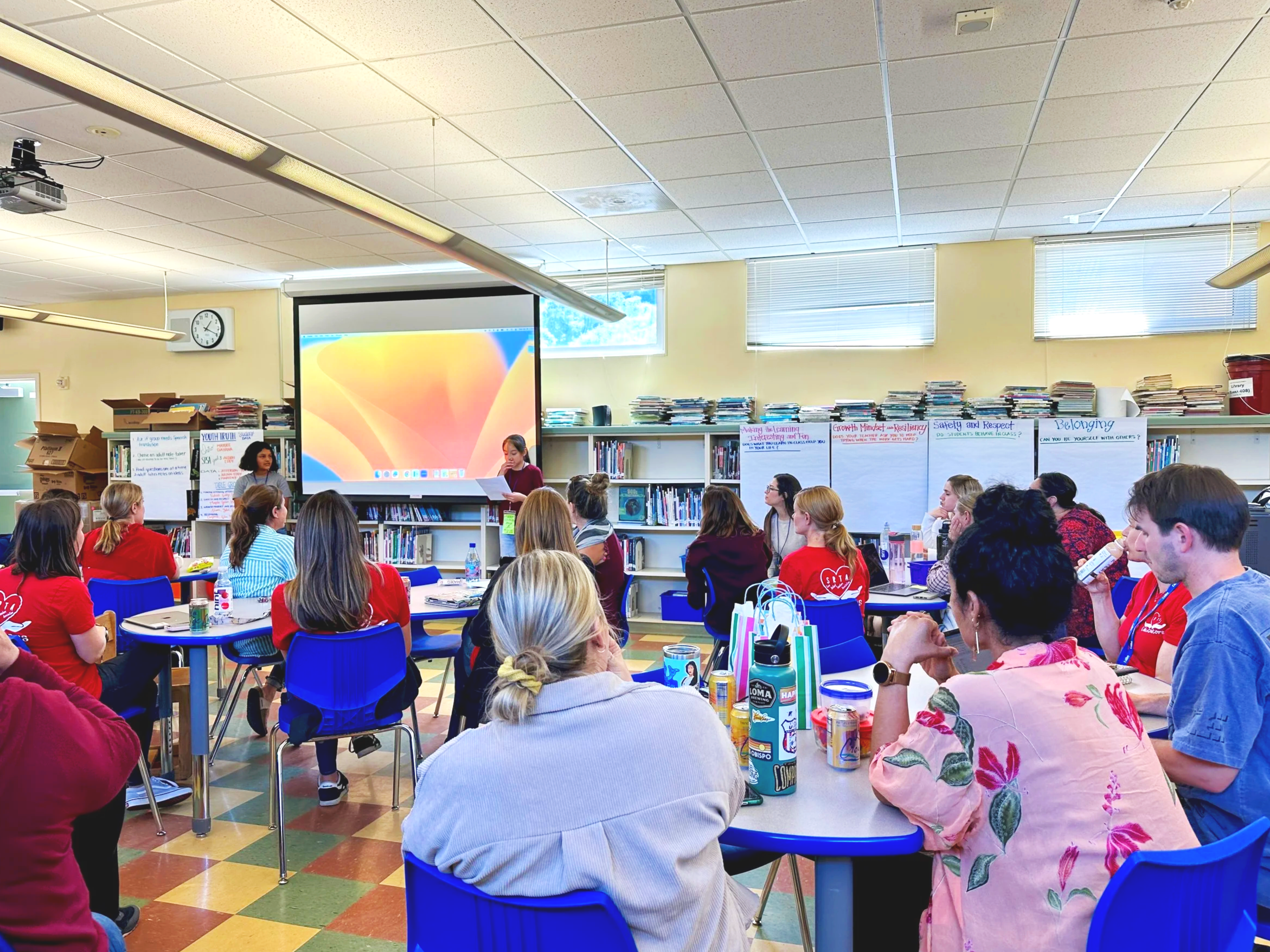 A school library with adults sitting around tables. There are bookshelves and posters on the walls, and a large projection screen at the front. Two adults are presenting at the front of the room.