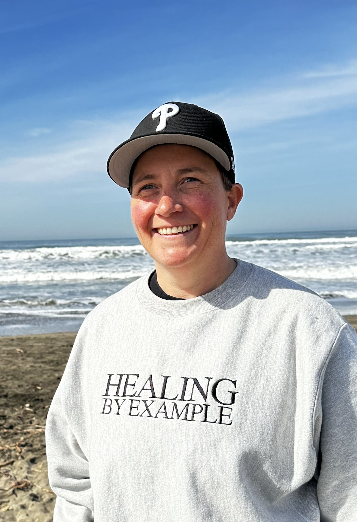 A woman with short brown hair wearing a black baseball hat and a grey sweatshirt with the words "Healing by Example" on the front. There are sand and the ocean in the background. 