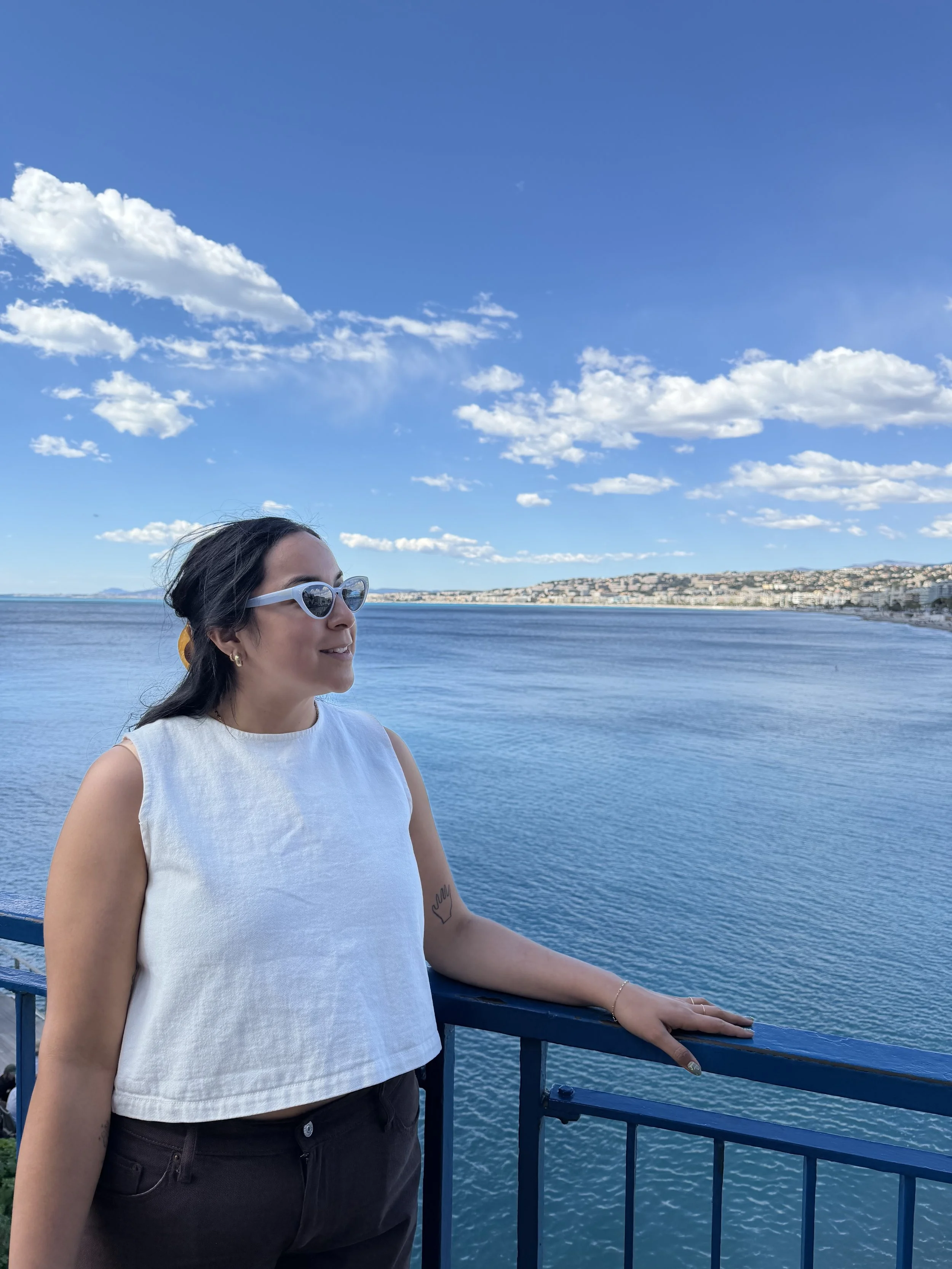 A woman with long dark black hair and dark brown skin, wearing white sunglasses, a white tank top, and black pants. There is water and a blue sky in the background.