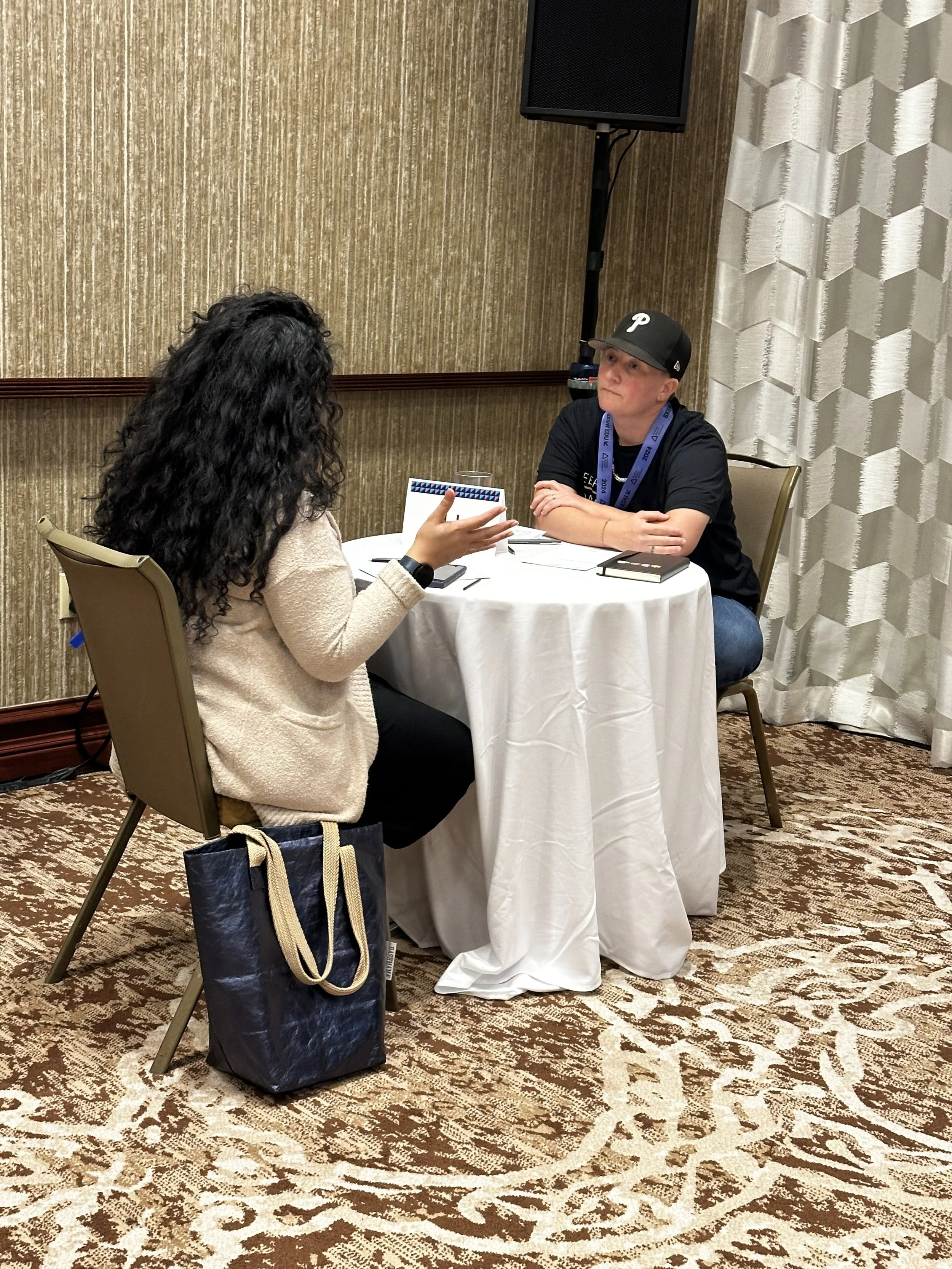 A person with curly black hair sitting at a round table, talking to a person wearing a black baseball cap and black T-shirt at a conference or seminar in a room with beige patterned carpet and textured beige walls.