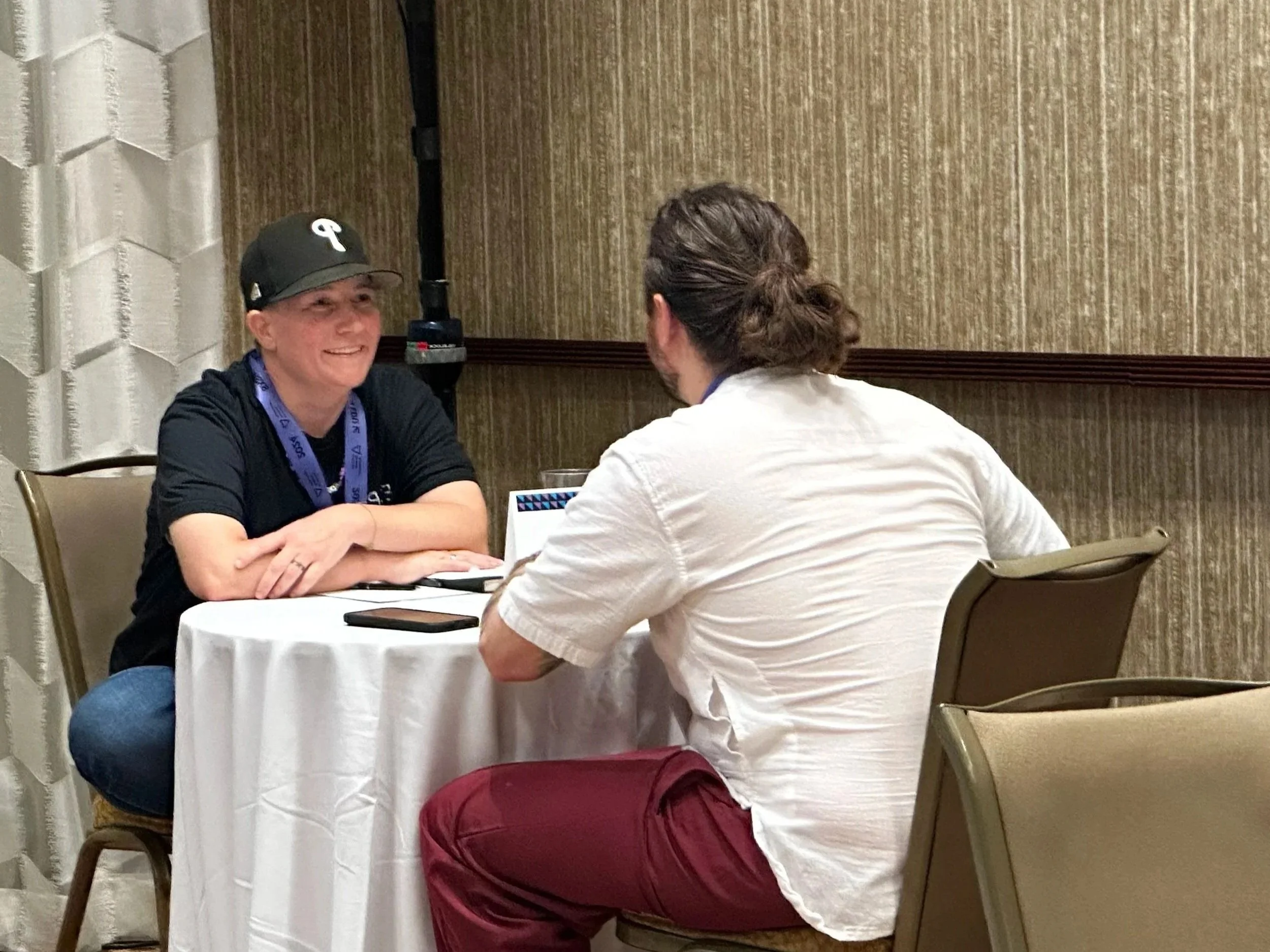 Two people sit across from each other at a round table, engaged in conversation inside a room with beige textured walls. The person on the left is smiling, wearing a black shirt .The person on the right facing away from the camera.