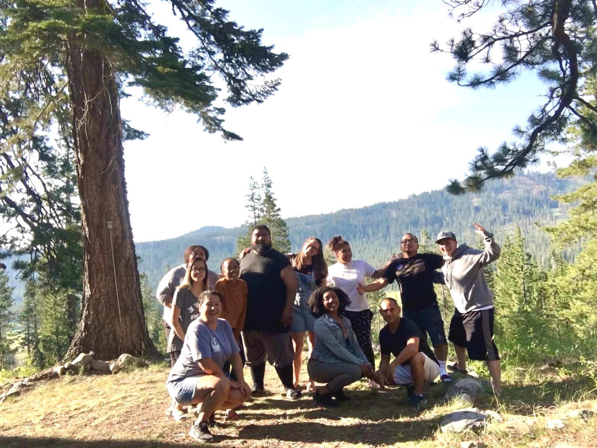 A group of people outdoors in a forested area with mountains and trees in the background, posing for a photo on a sunny day.