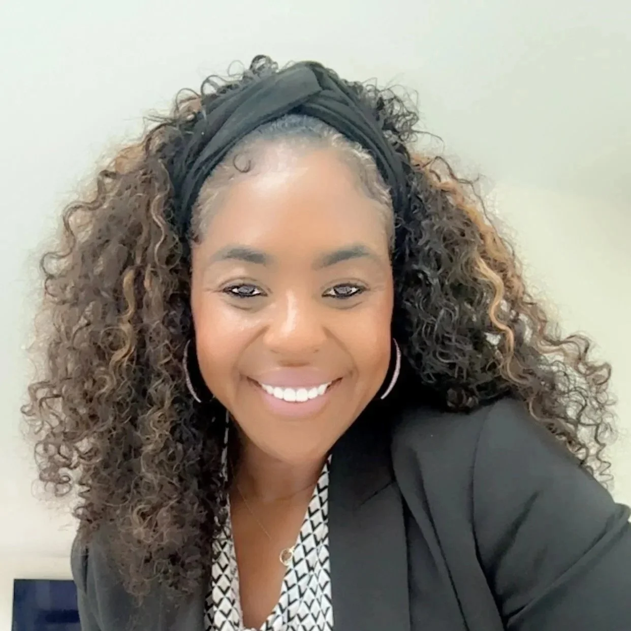 A woman with long dark curly hair a black headband, silver hoop earrings and a black blazer.