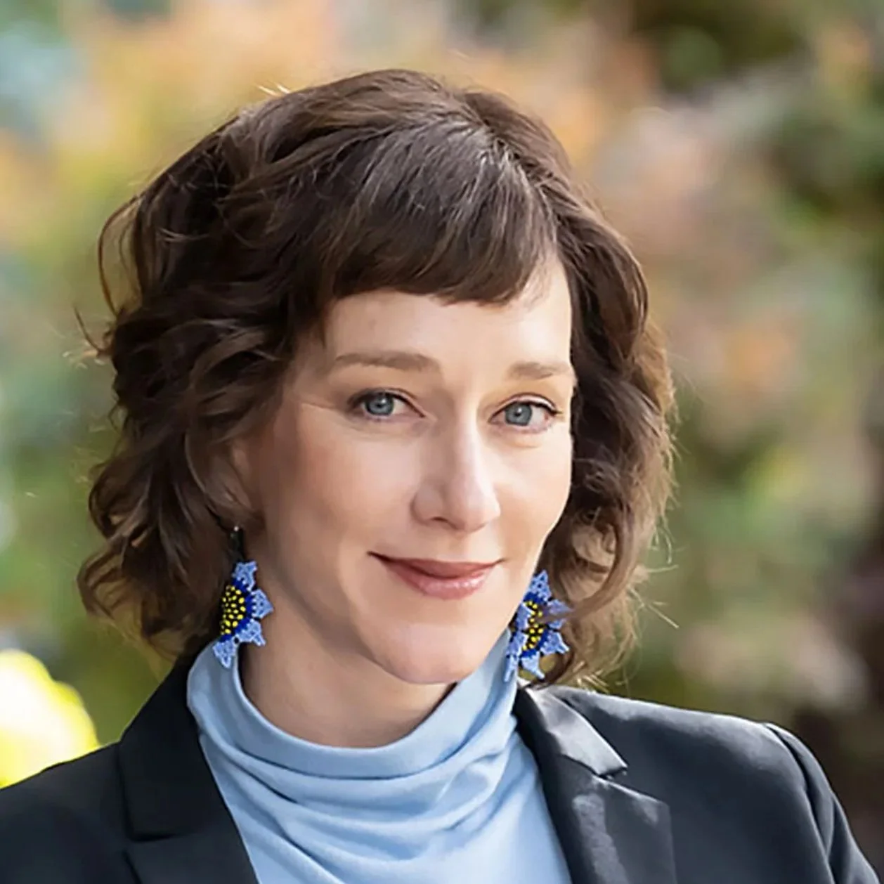 A woman with dark brown curly hair with blue flower earrings, a blue turtleneck, and trees in the background.