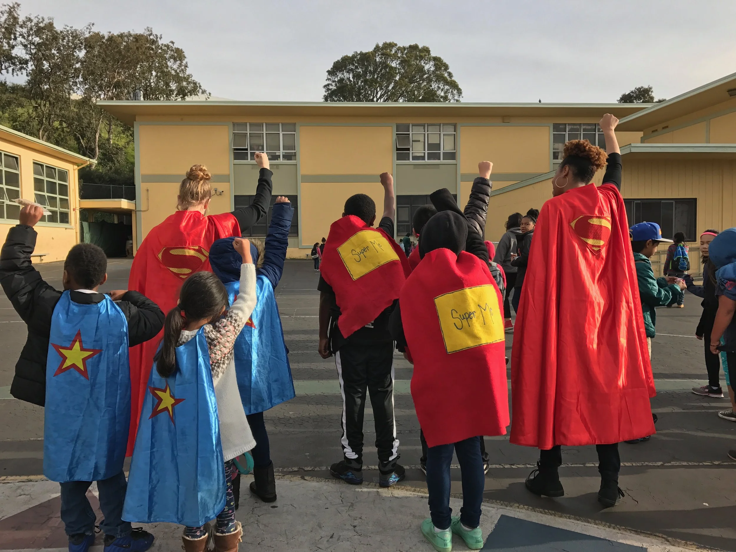 A group of children and adults wearing superhero capes, some with raised fists, gathered outdoors in front of a yellow school with trees in the background.