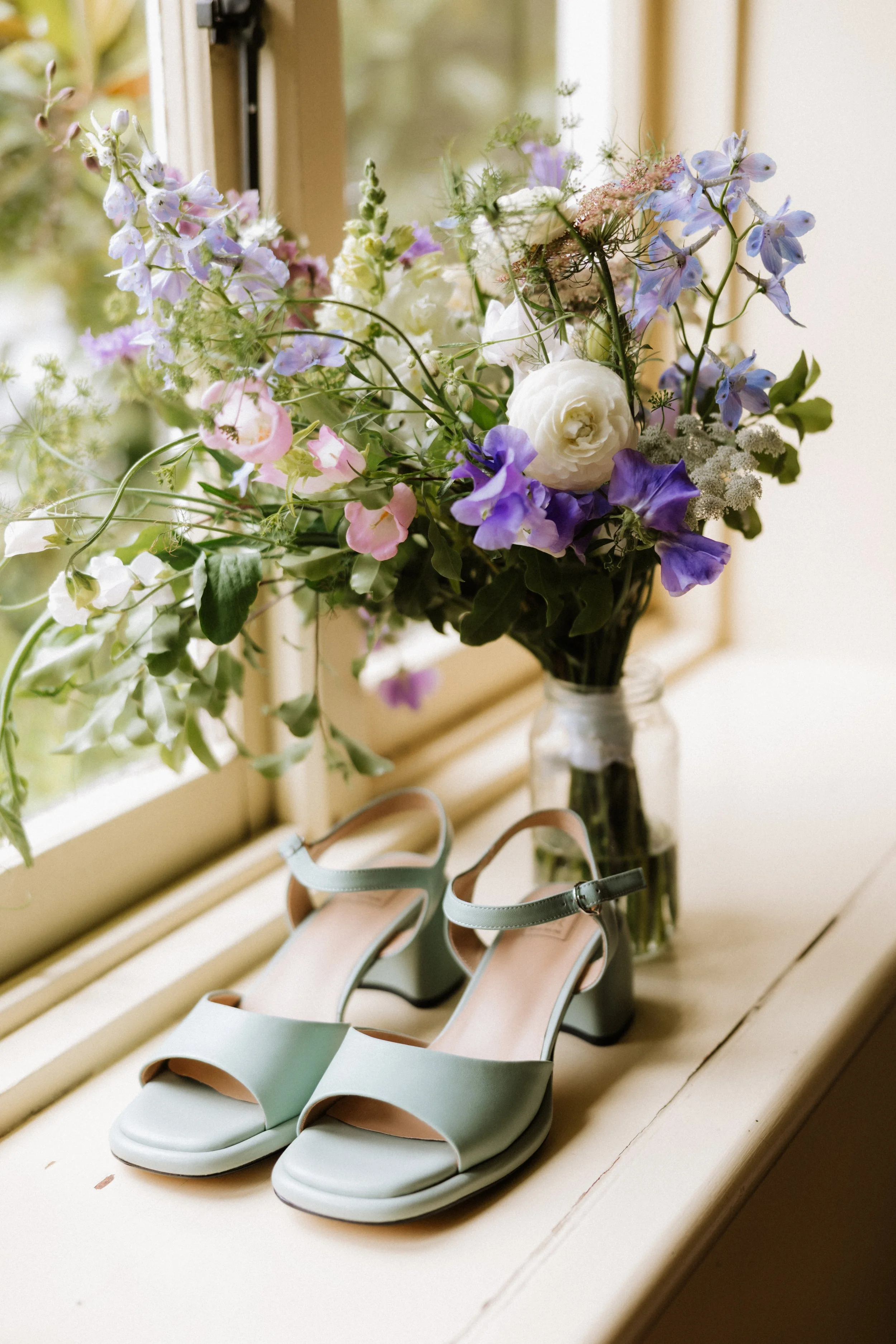 A pair of light green heeled sandals and a bouquet of purple, white, and pink flowers in a glass jar on a windowsill.