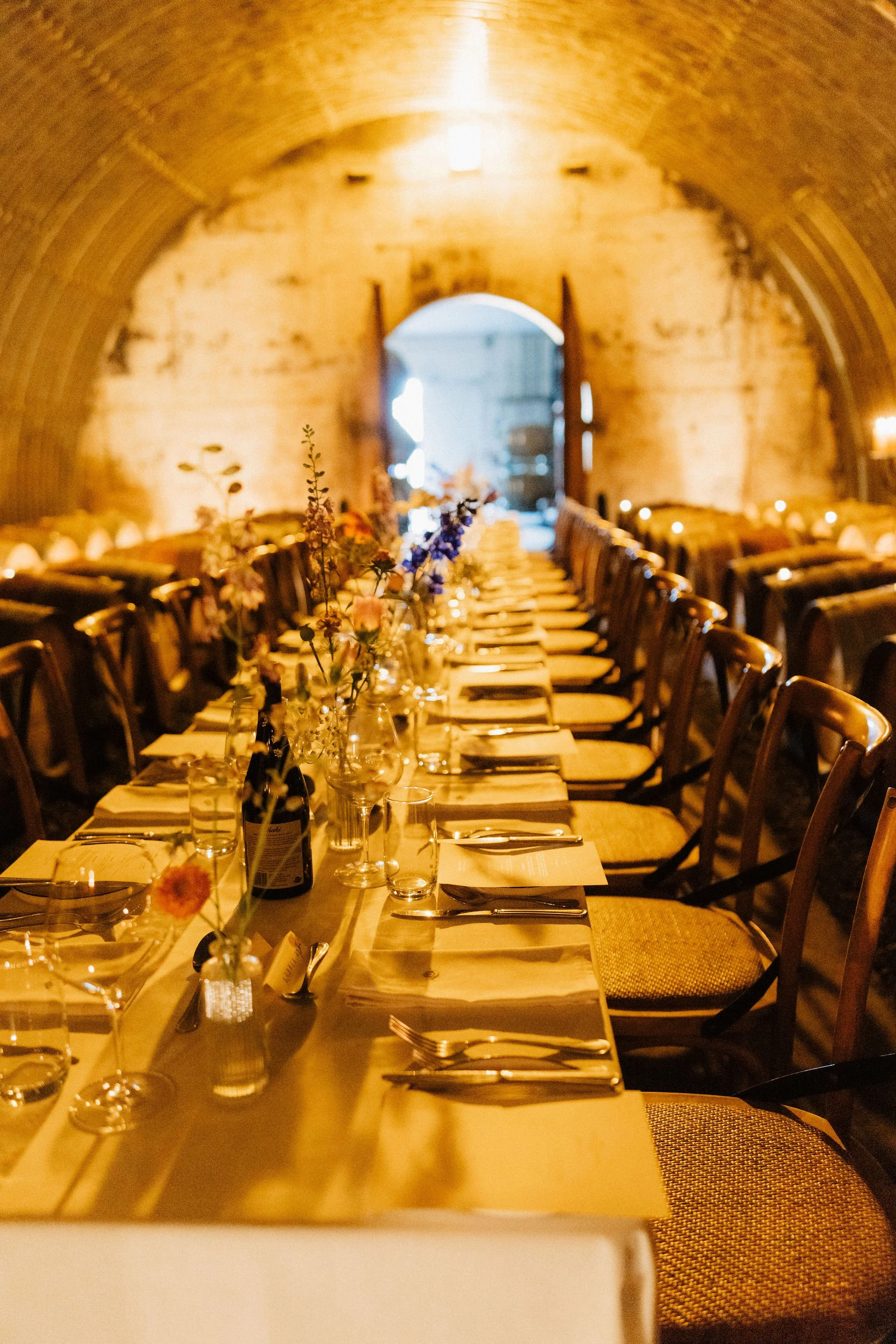 A long banquet table set for a formal dinner in a rustic stone-walled room with an arched ceiling, decorated with flowers, glassware, and cutlery.