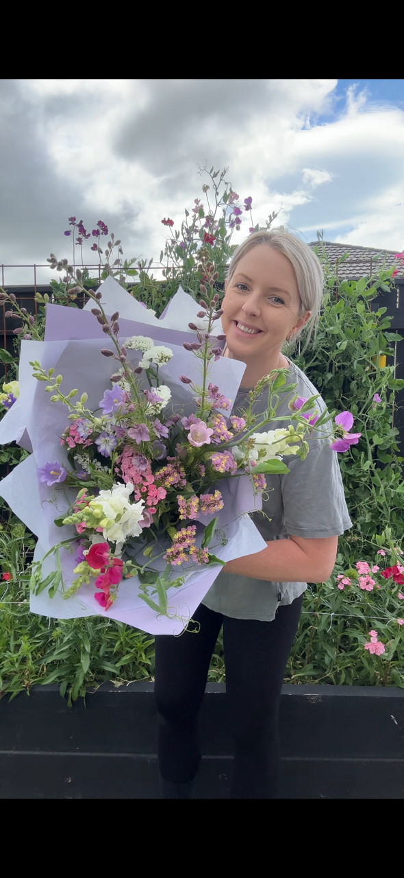 Woman holding a large bouquet of colorful flowers in a garden with green plants and flowers, cloudy sky in the background.