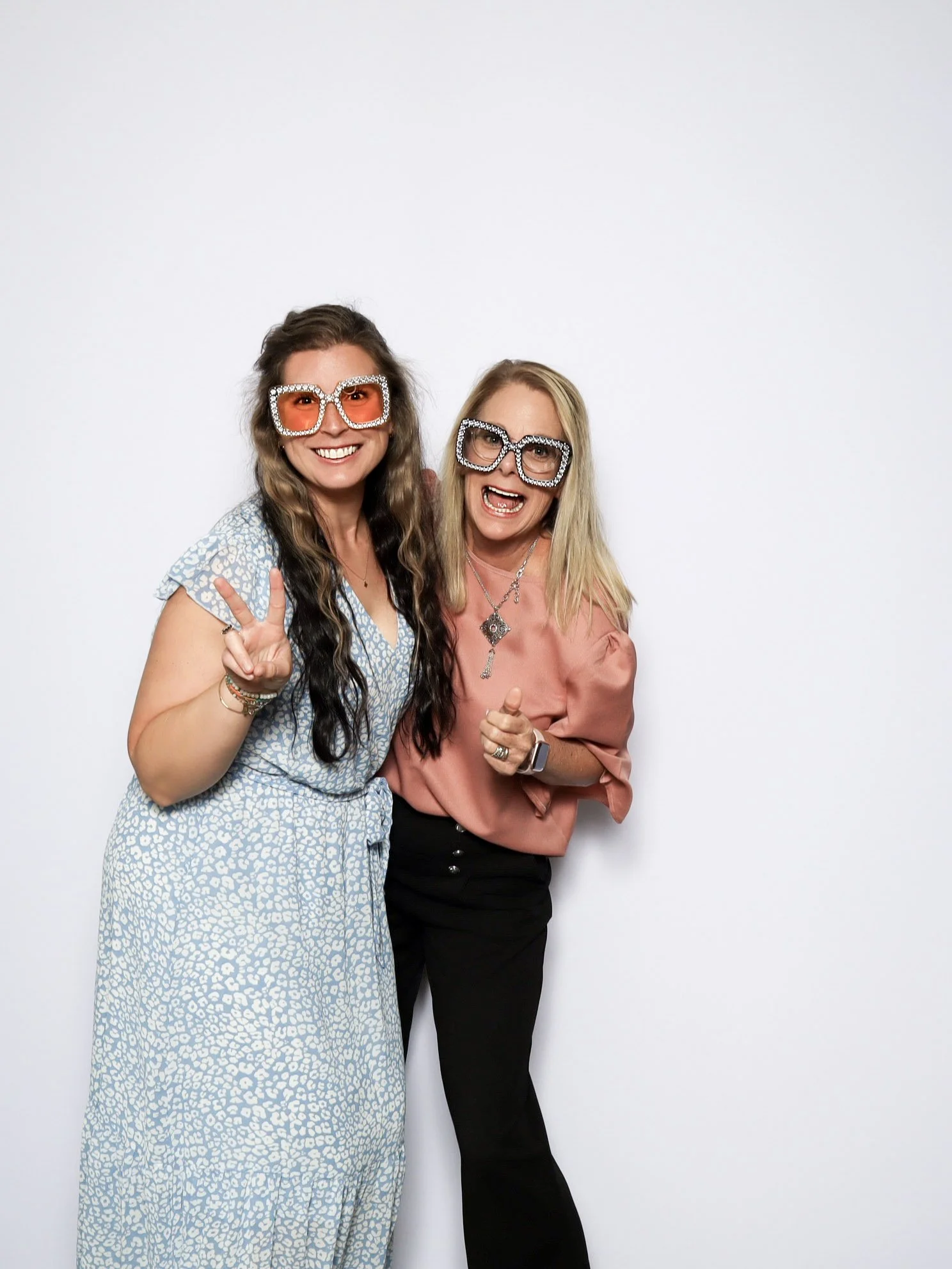 Two women in colorful retro glasses pose and smile for a photo against a plain white background.