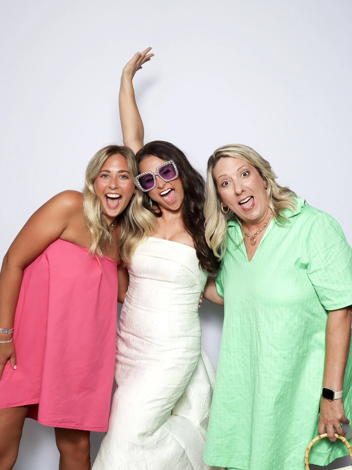 Three women celebrating together, one in a pink dress, one in a white dress with purple sunglasses, and one in a green dress, against a white background.