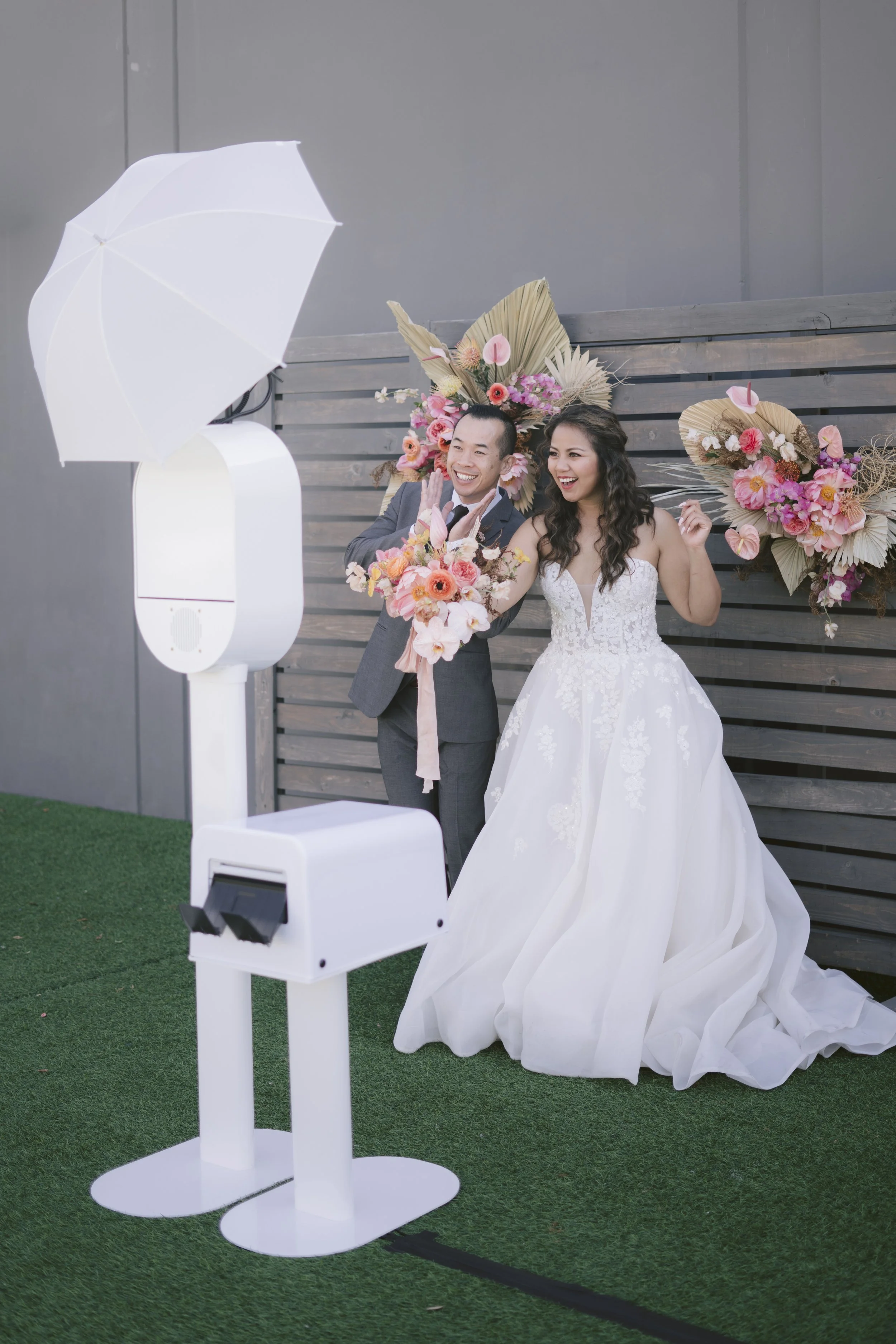 Bride and groom smiling while posing at a PRO photo booth setup with prints at a wedding event. Bride in a white gown holding flowers, groom in a gray suit, floral backdrop style, white umbrella reflector lighting. Keywords: PRO photo booth rental