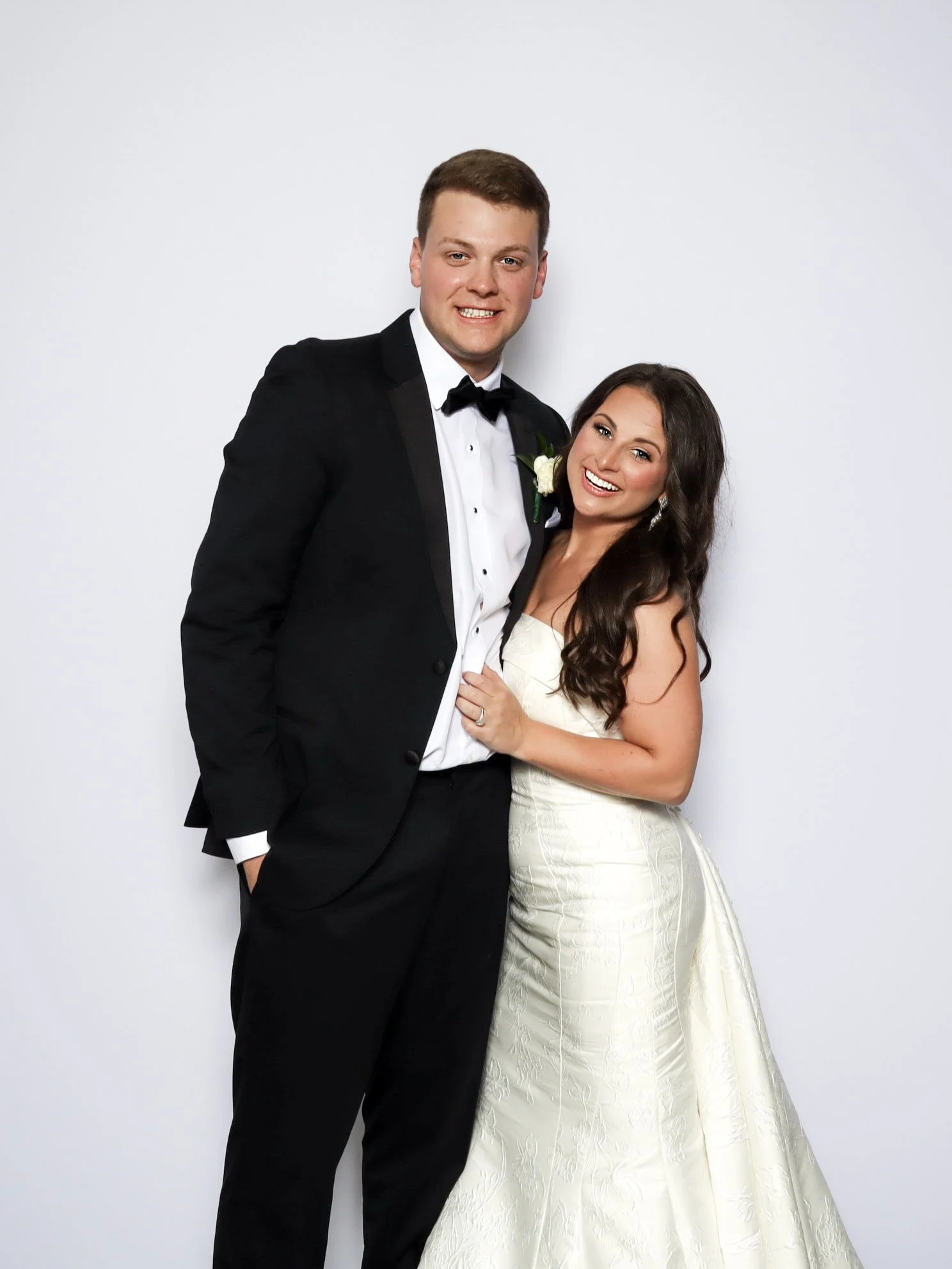 A bride and groom in wedding attire standing together and smiling in front of a plain white background.