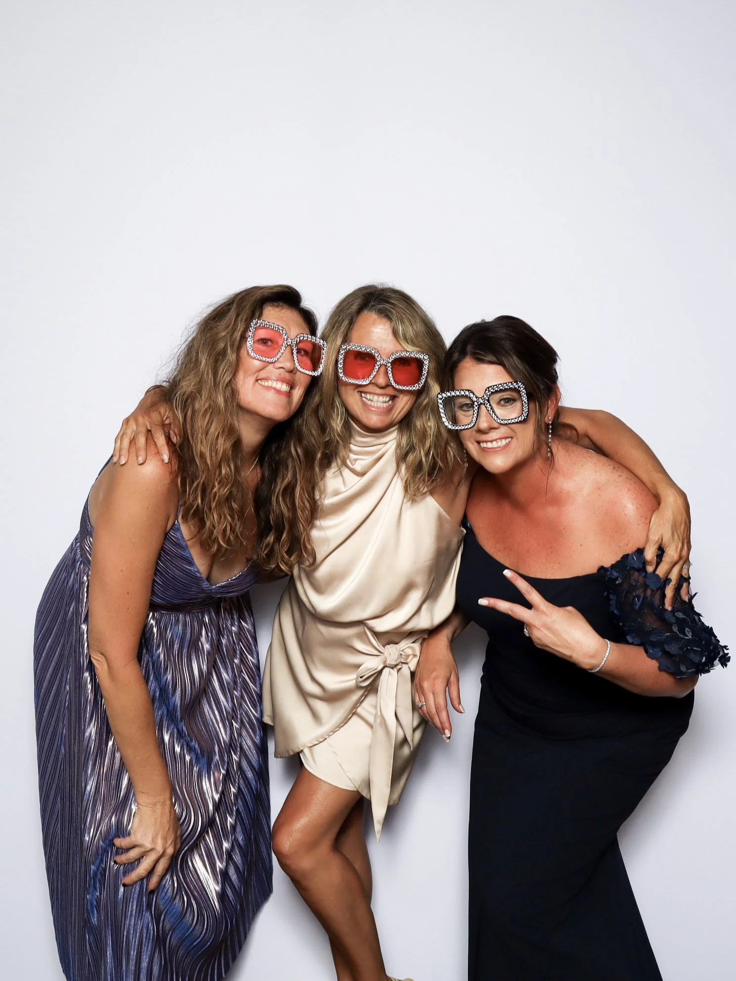 Three women smiling and posing together, wearing large, sparkly sunglasses, with arms around each other, standing against a plain white background.